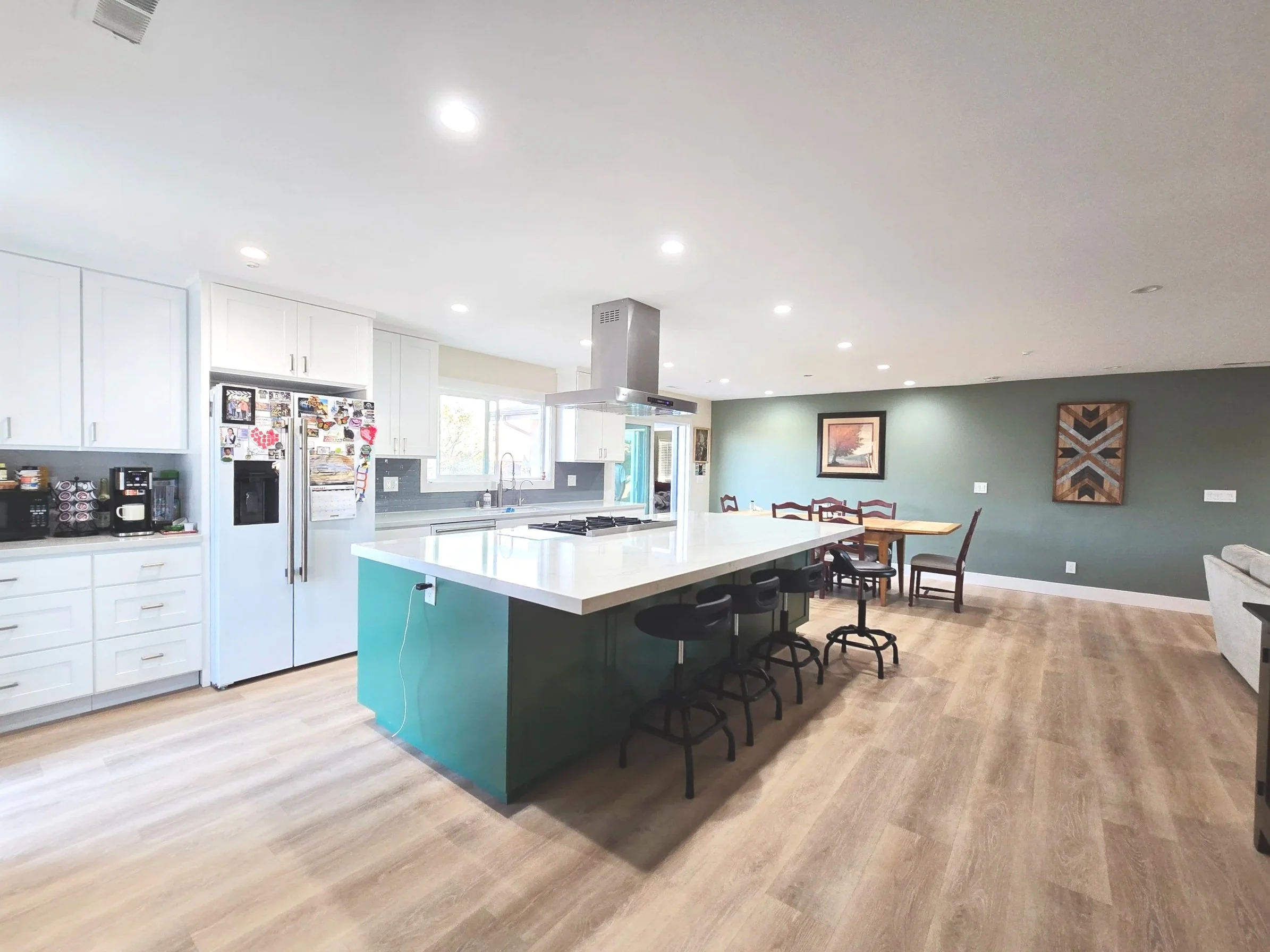 A modern open-concept kitchen and dining area with white cabinets, a kitchen island, and a dining table with chairs on wood flooring. There is a green accent wall with framed artwork.