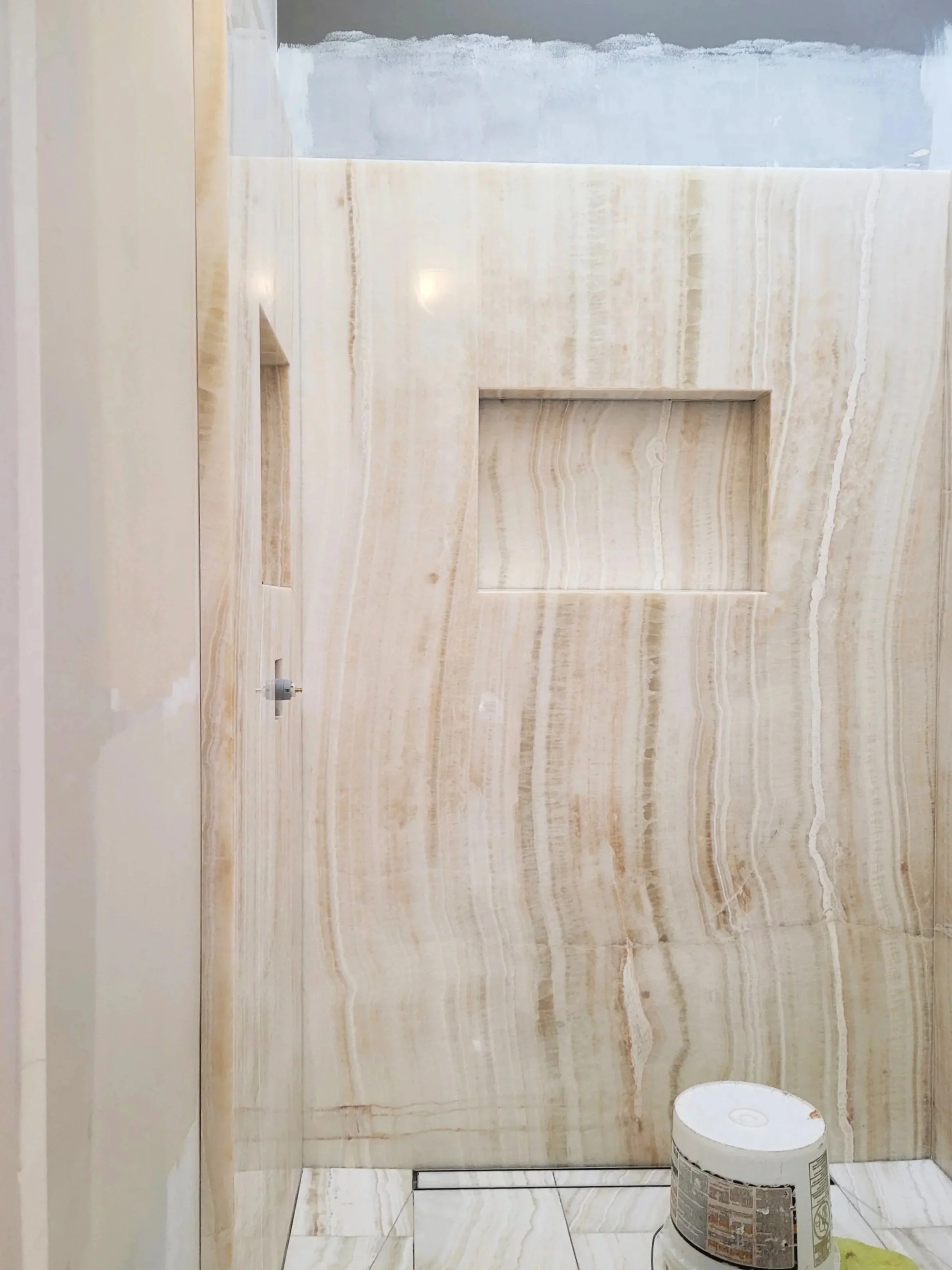 A bathroom marble wall with a small recessed shelf, a bucket of construction material, and a bucket of paint or plaster, indicating ongoing renovations.