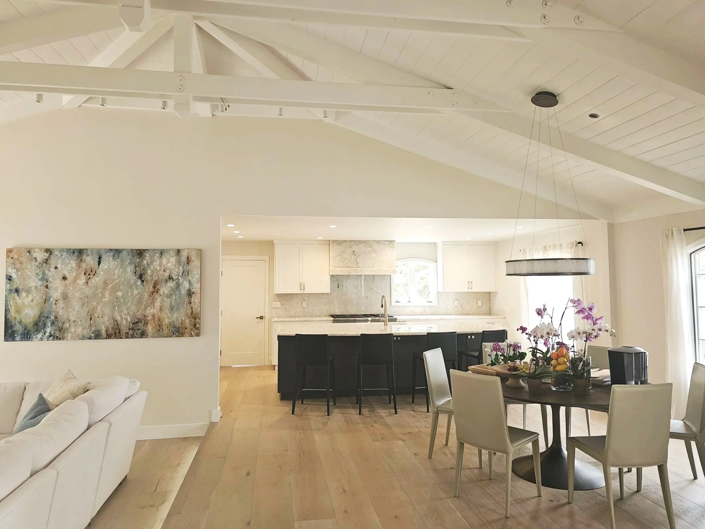 Open-concept living and dining area with white walls, a wooden floor, a white couch, a large abstract painting, and a dining table with black and white chairs. The kitchen has white cabinets and marble countertops, with a pendant light over the dining table and purple orchids on the table.