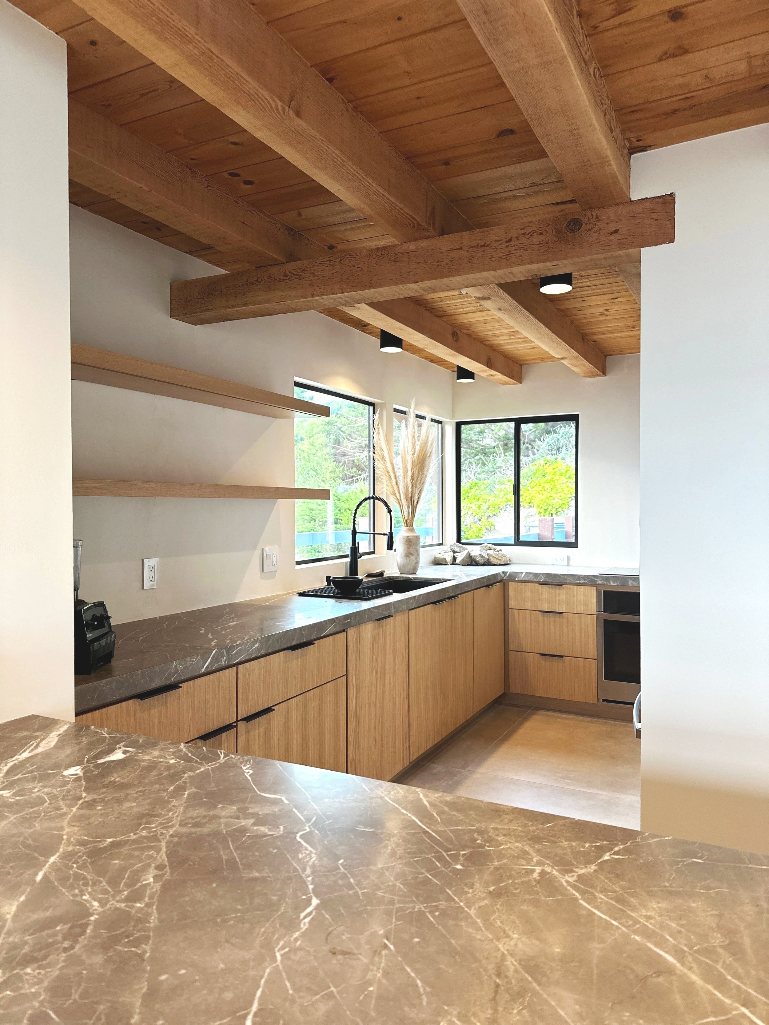Modern kitchen with wooden cabinets, marble countertops, black sink with a black faucet, and large windows showing greenery outside. Wooden ceiling beams and black ceiling lights.