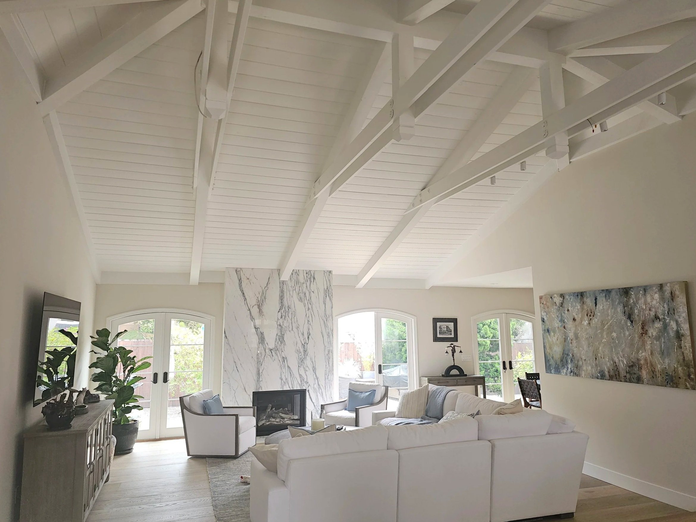 Living room with white vaulted ceiling with exposed beams, large windows, white sofa, and artwork on the wall.