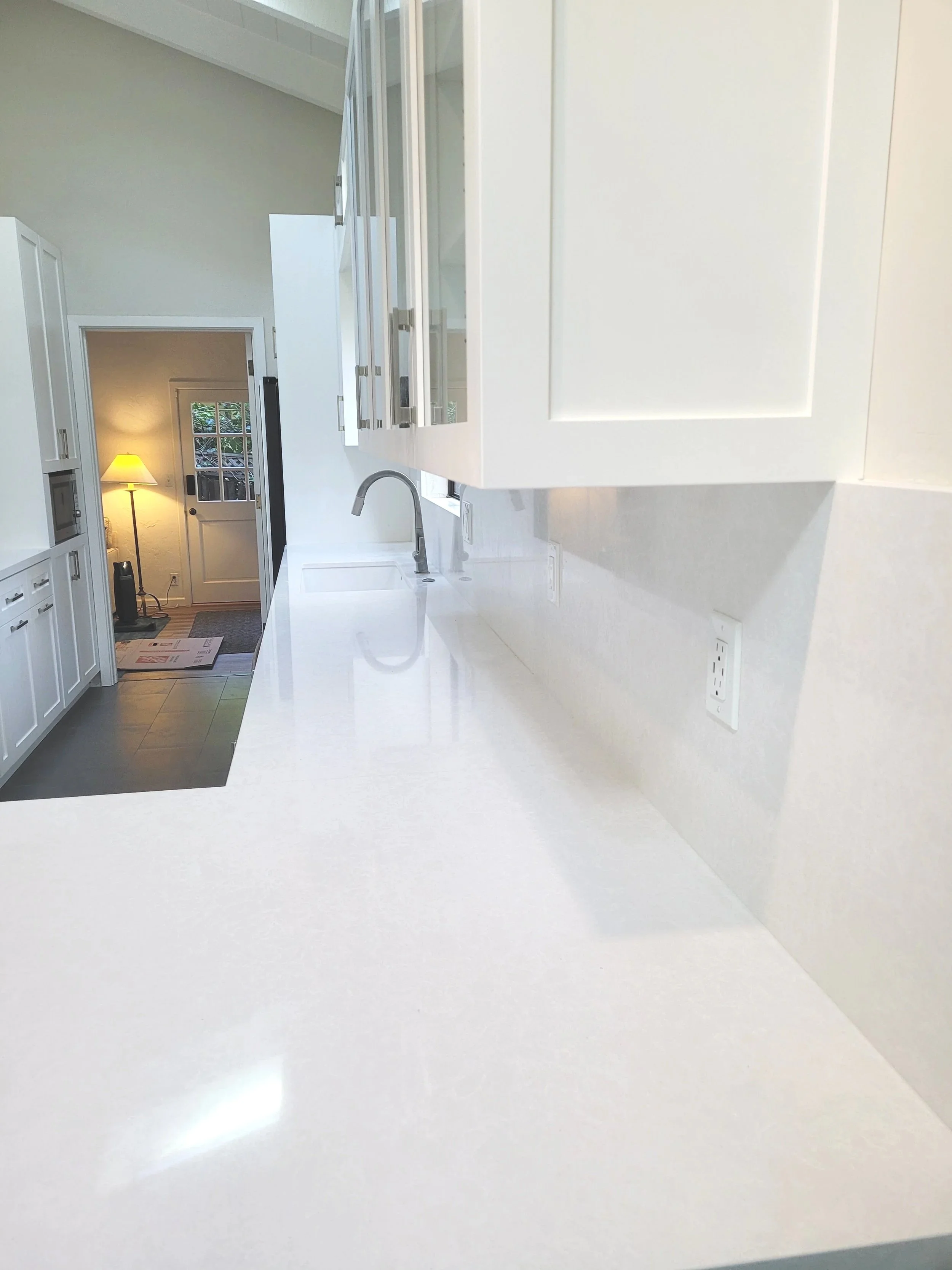The image shows a clean, white kitchen counter with a sink and a modern faucet, overlooking a bright, airy room with white cabinets and a window. In the background, a door leads to an entryway with a small lamp, a door with windows, and some scattered papers and a rug.