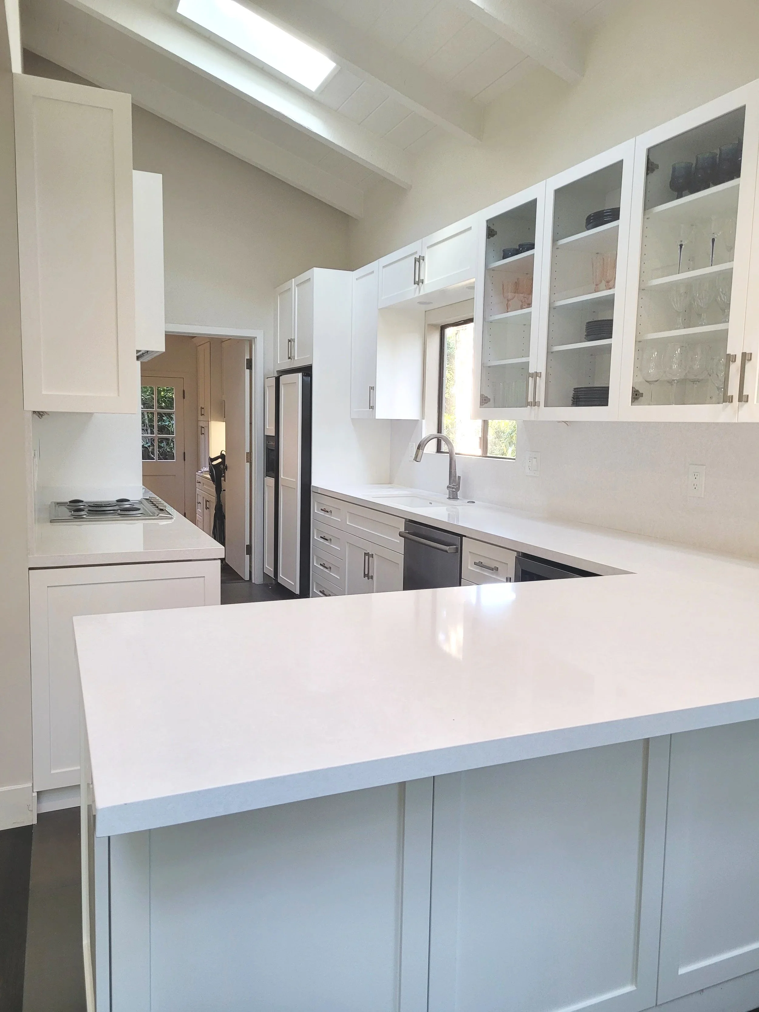 Modern white kitchen with open upper cabinets displaying dishes and glasses, a stainless steel refrigerator, a window above the sink, and a kitchen island with a white countertop.