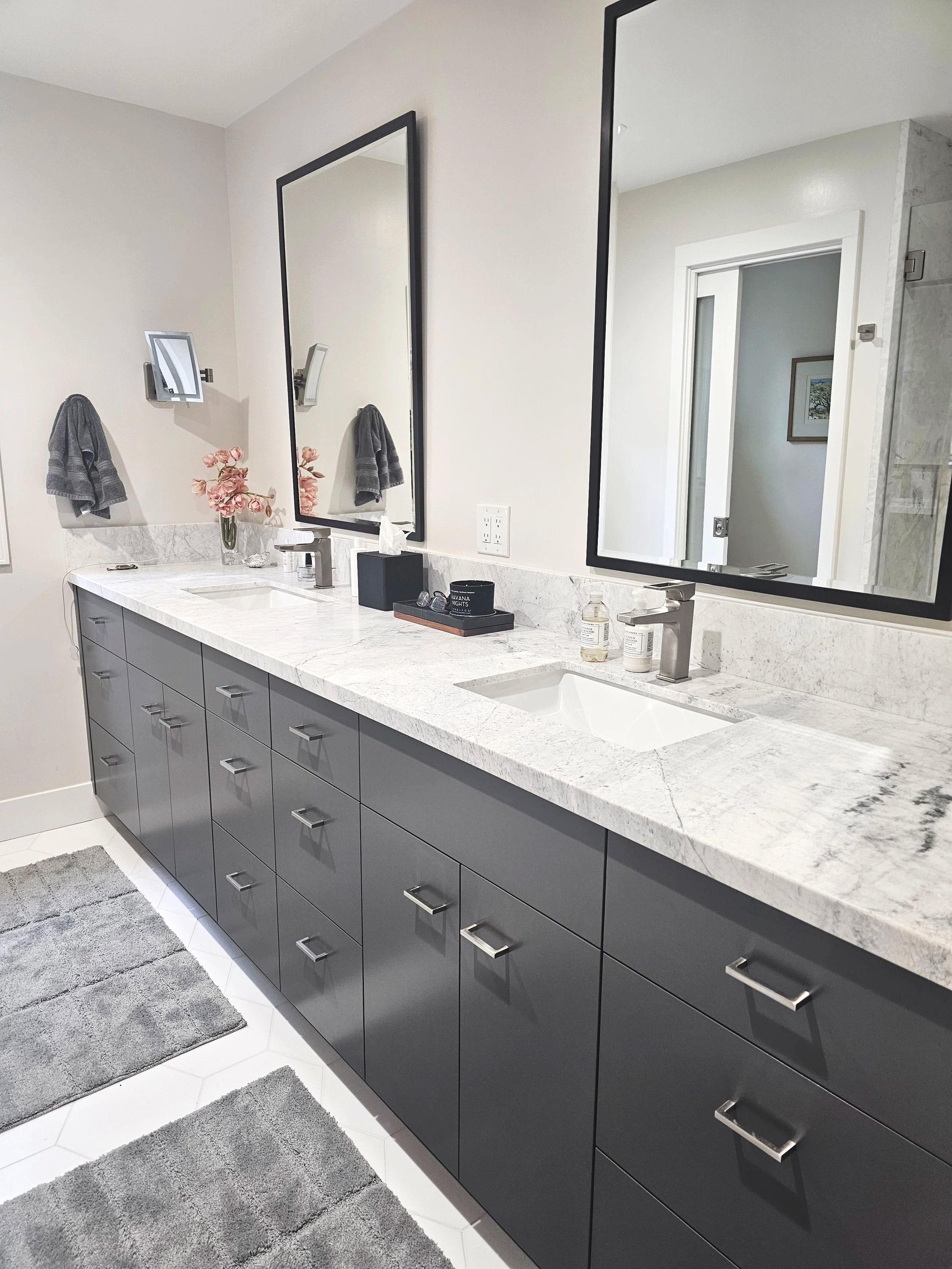 Modern bathroom with double vanity with gray cabinets, white marble countertop, two rectangular mirrors, two faucets, a pink flower vase, and various toiletries; gray towels hanging on the wall; small framed art in the background; hexagonal floor tiles and gray rugs.