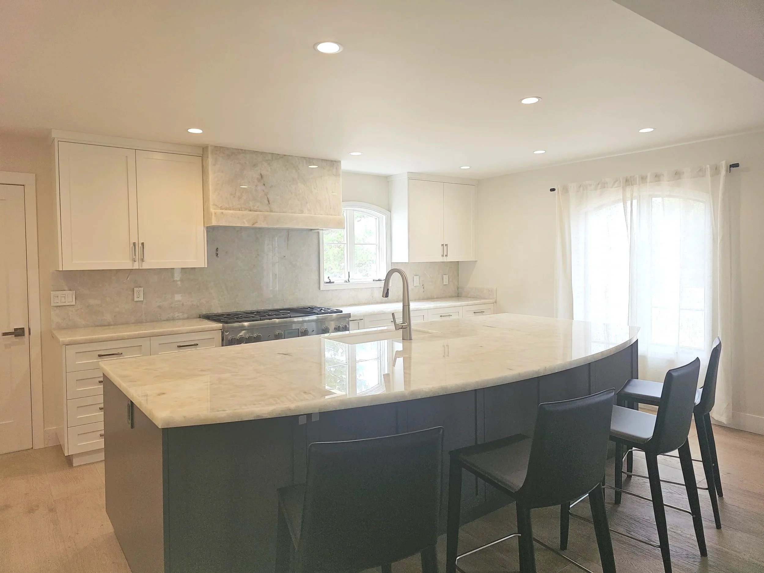 Modern kitchen with white cabinetry, a marble backsplash, large kitchen island with marble top, black chairs, stainless steel stove, a window, and curtains.