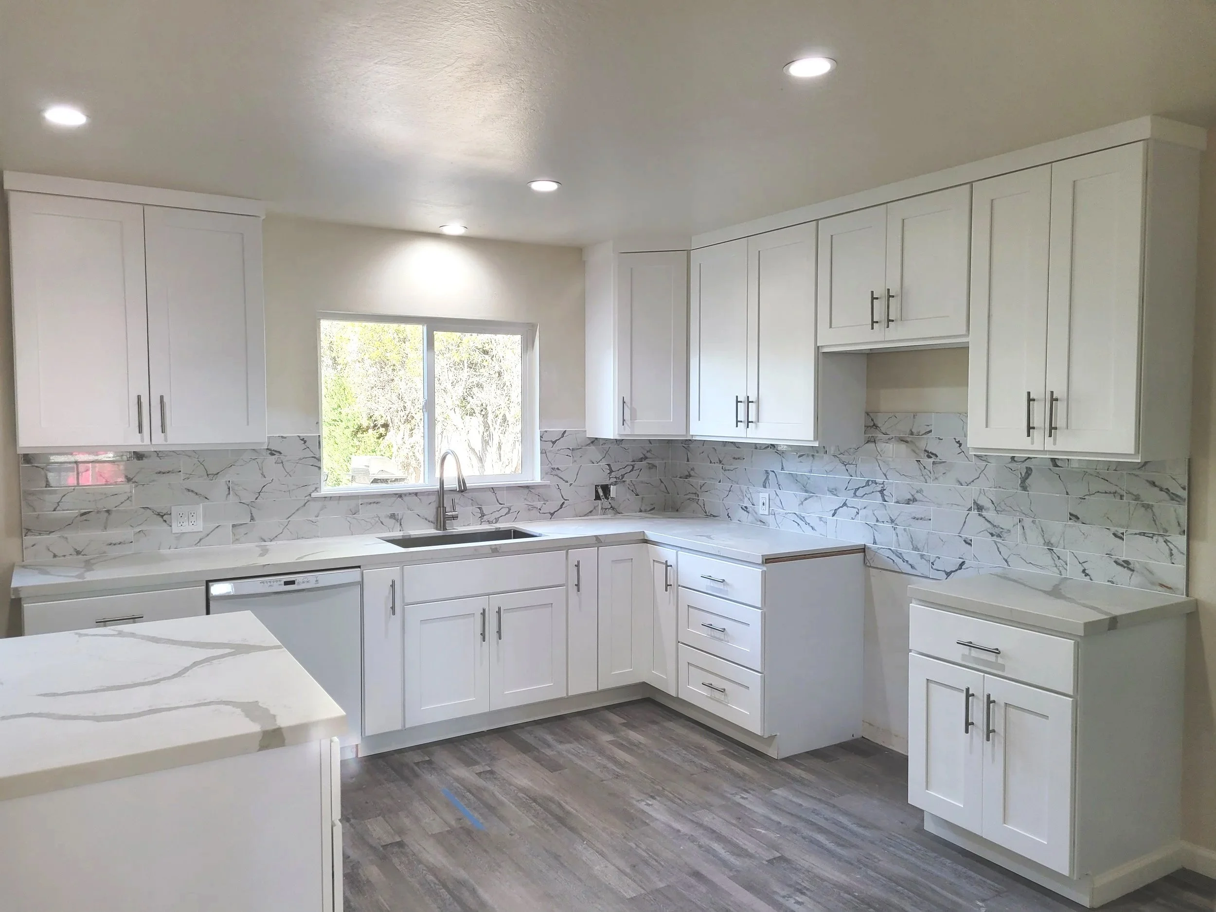 A modern kitchen with white cabinets, marble backsplash, and a large window above the sink, with recessed lighting in the ceiling.