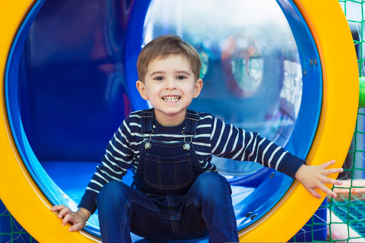 A young boy with physical disabilities smiling and sitting inside a colorful play tunnel at a sensory-friendly, inclusive indoor playground in Orange County, CA.