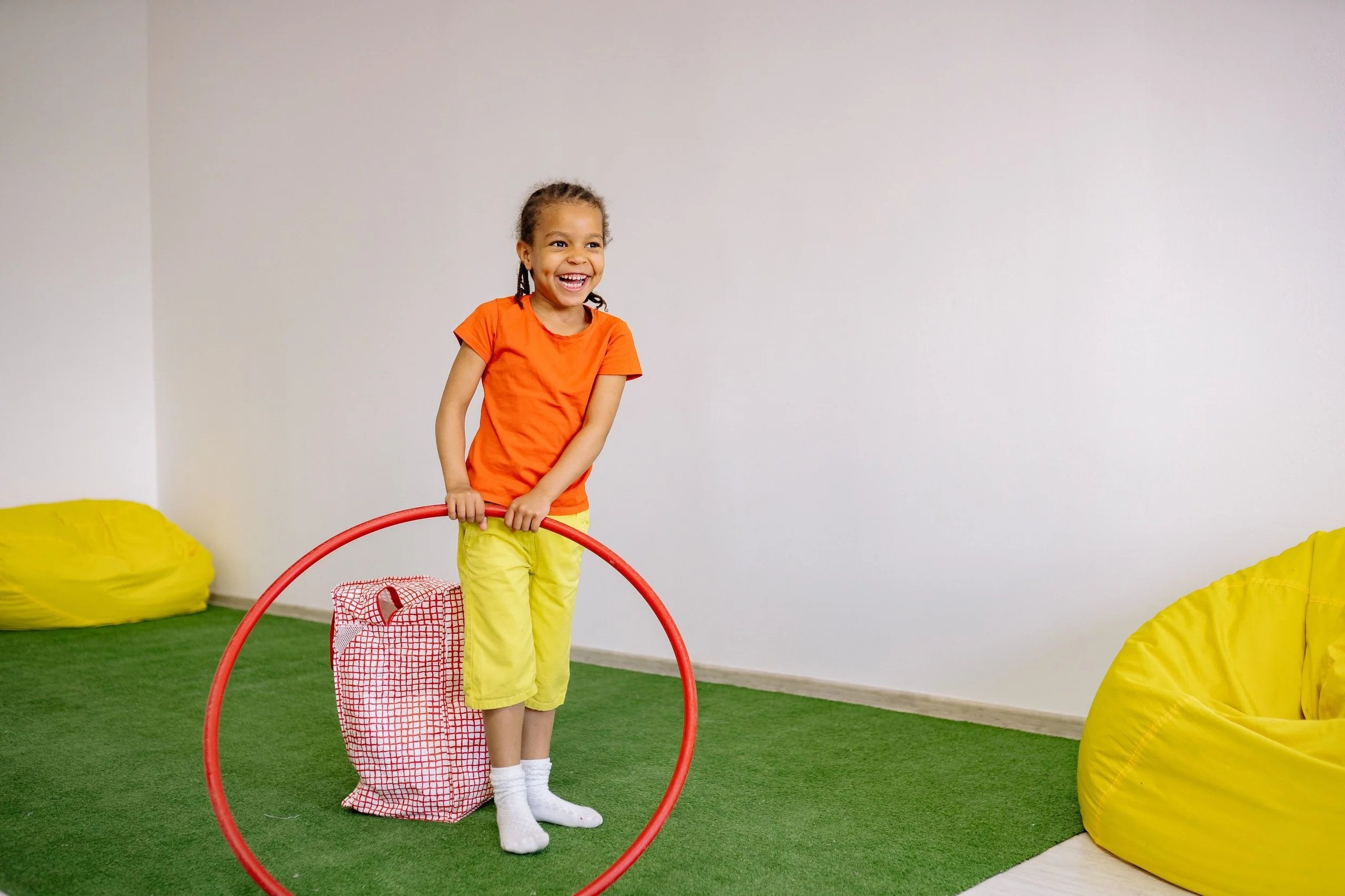 A young girl smiling and playing with a red hula hoop indoors at a sensory-friendly play gym in Orange County, CA