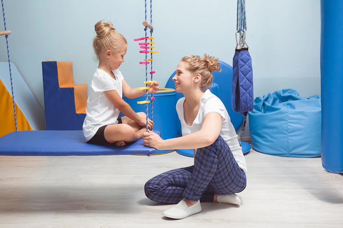 A young girl sitting on a swing with a physical therapist kneeling in front of her, smiling. The physical therapist is using space at an indoor, sensory gym in Orange County, CA