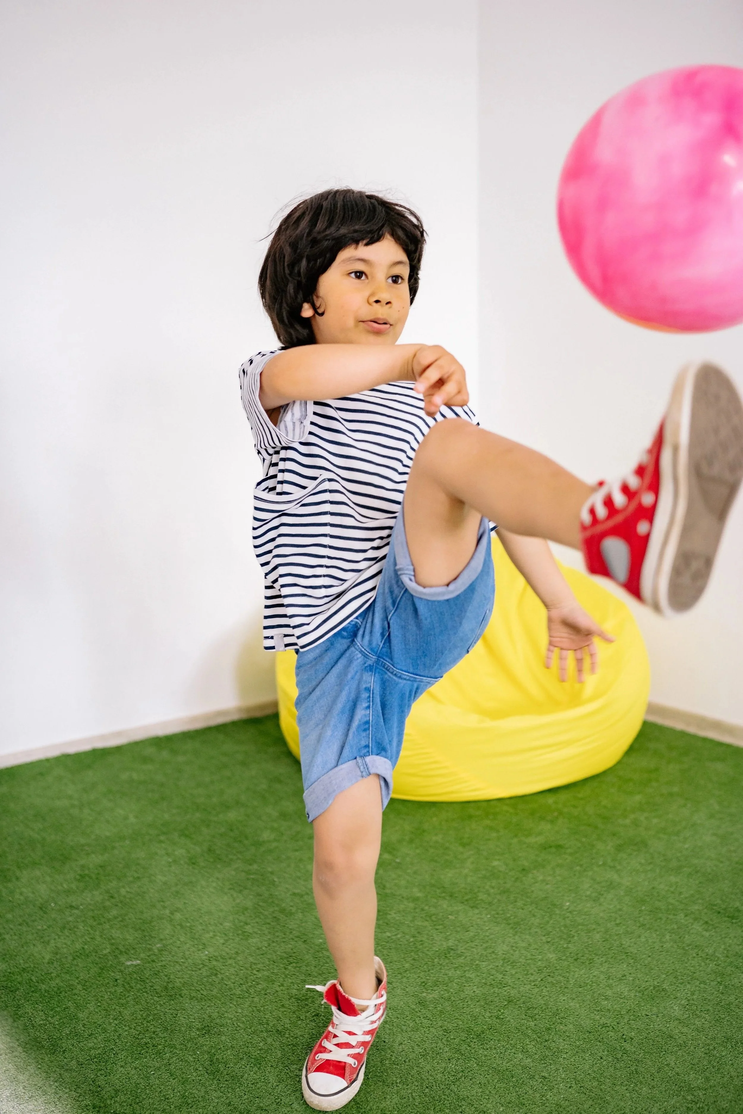 A neurodivergent boy wearing a striped t-shirt and red sneakers kicking a ball during sensory play at an indoor, inclusive play gym in Orange County, CA