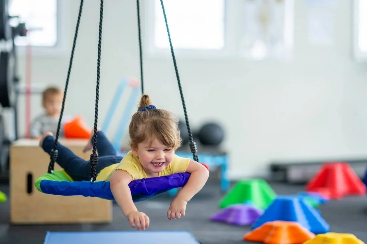 Young girl with autism swinging on a sensory swing with other climbing equipment and children in the background, playing at an indoor sensory gym in Orange County, CA