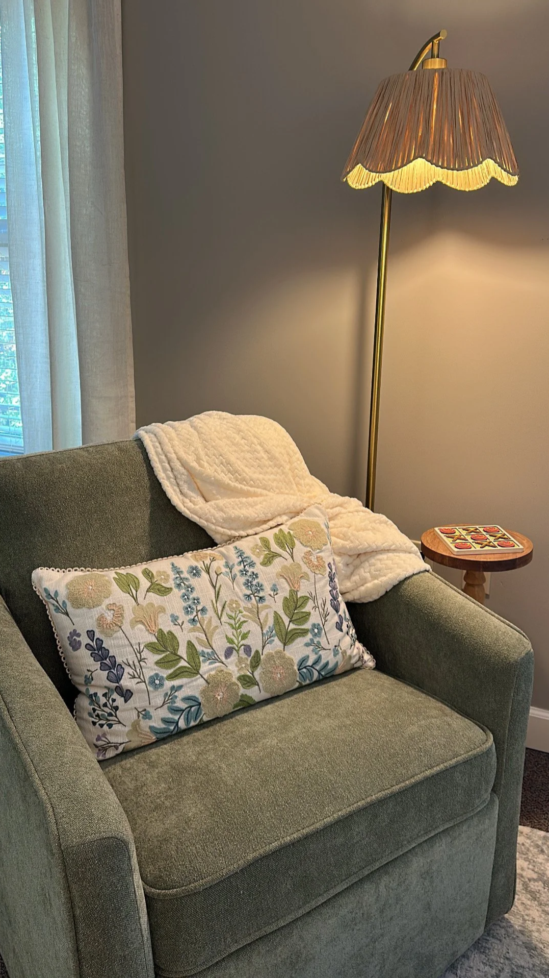 A living room corner featuring a green upholstered sofa with embroidered floral pillow and a cream blanket. There is a tall, gold floor lamp with a scalloped pink lampshade next to the sofa. A small round wooden side table with a Tic-Tac-Toe game is beside the lamp. A window with sheer curtains is in the background.