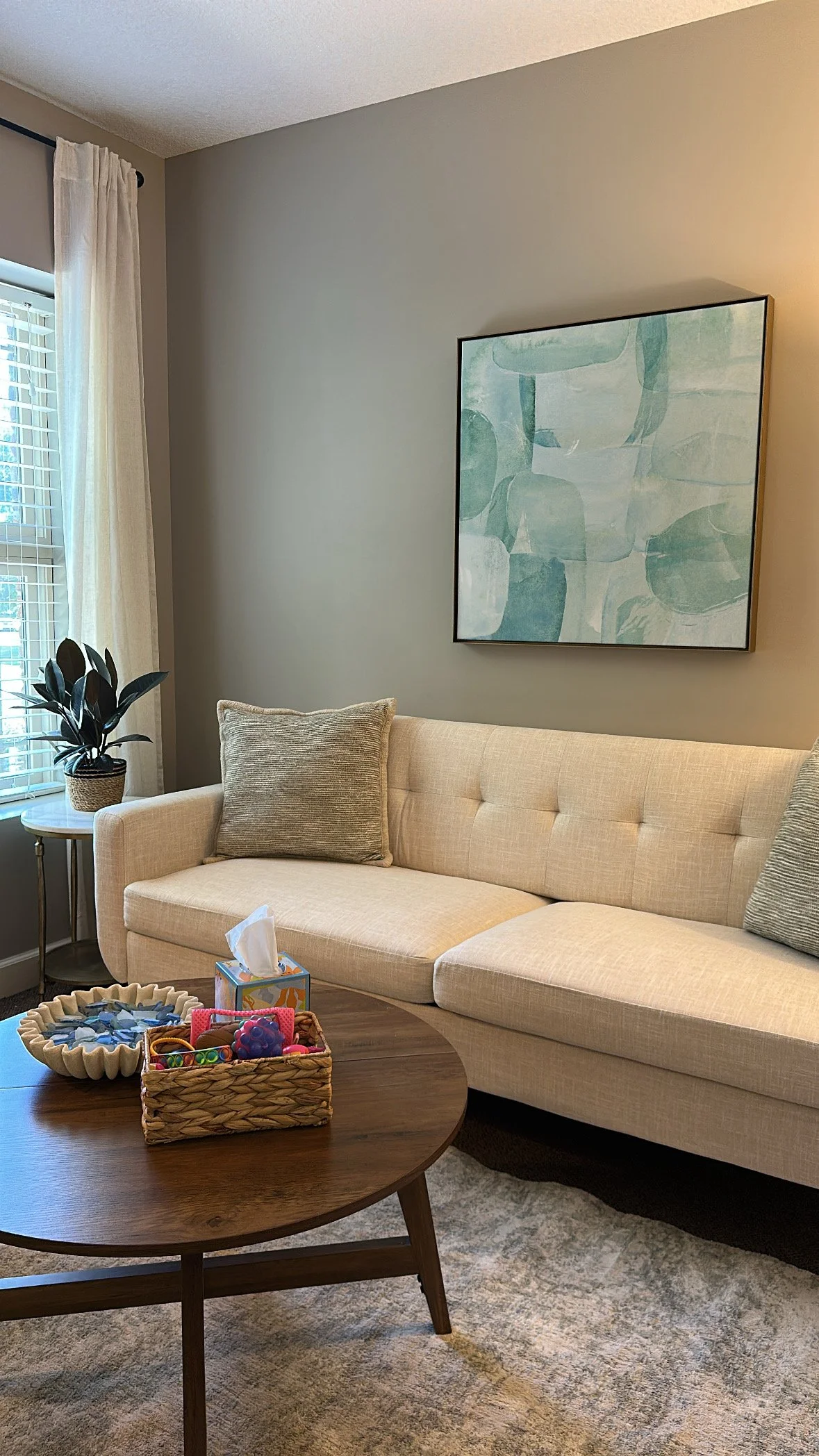 A living room with a beige sectional sofa, gray pillows, a wooden coffee table, a potted plant next to a window with white curtains, a colorful basket with toys, and an abstract painting on the wall.