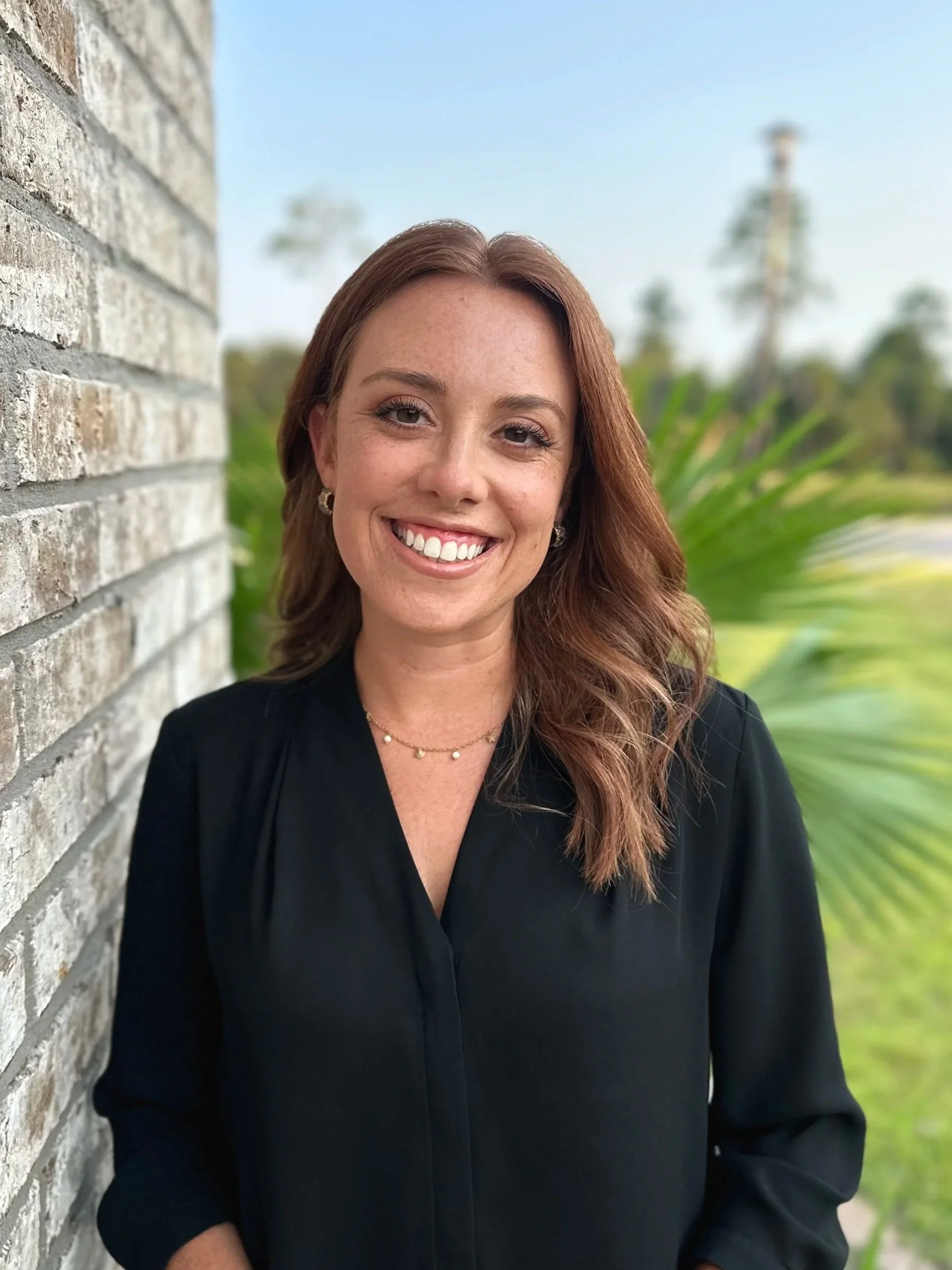 A woman with long, wavy red hair smiling and standing outdoors next to a gray brick wall, with greenery and trees in the background.