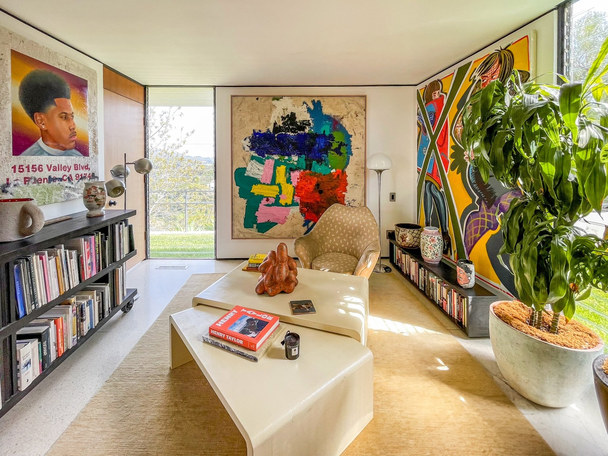 A cozy living room features abstract and portrait artwork on the walls, a small bookshelf, a beige patterned armchair, a white coffee table with books and decor, large potted plant, and a window with sunlight streaming in.