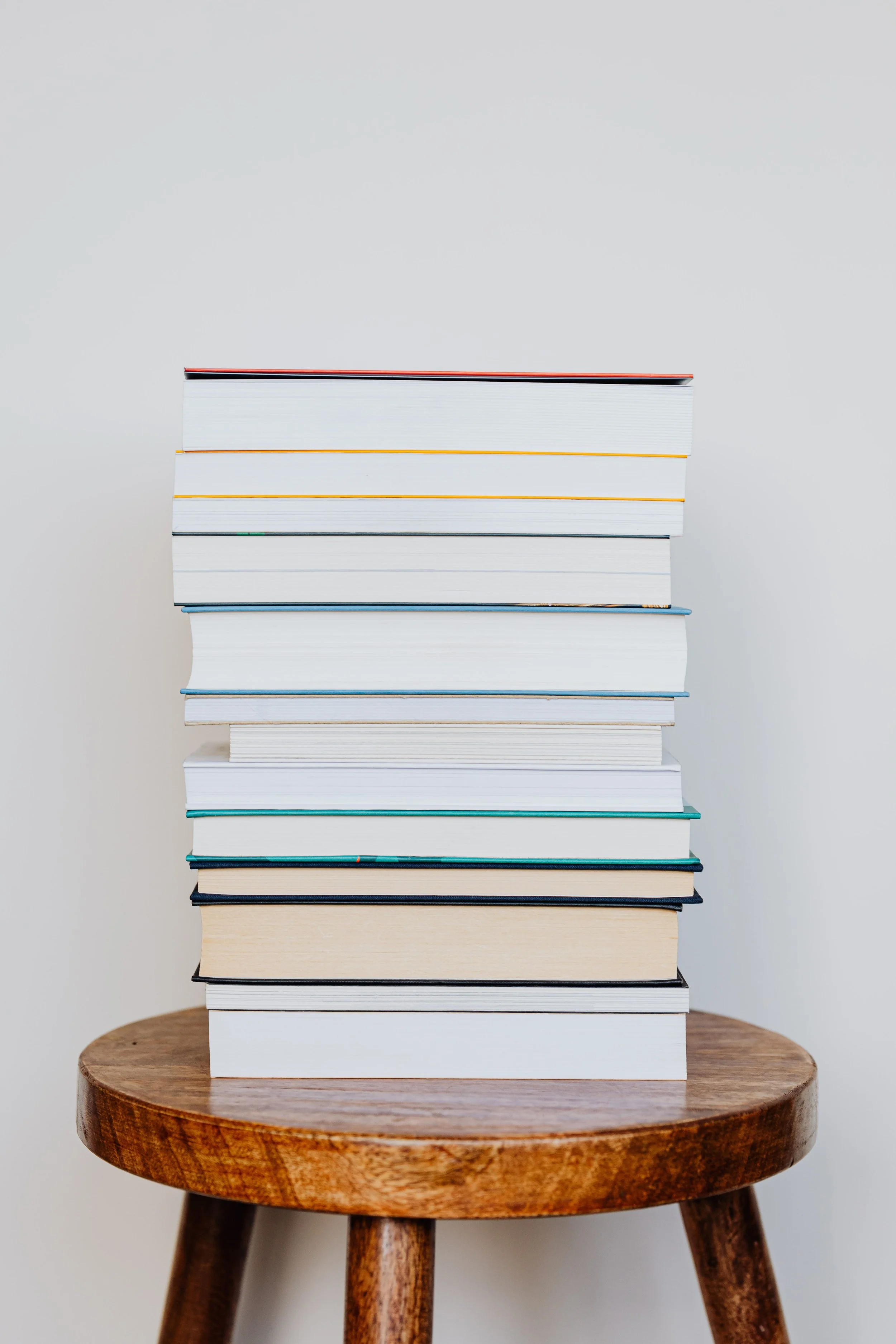Stack of white, black, and colored books placed on a wooden stool against a plain light background.