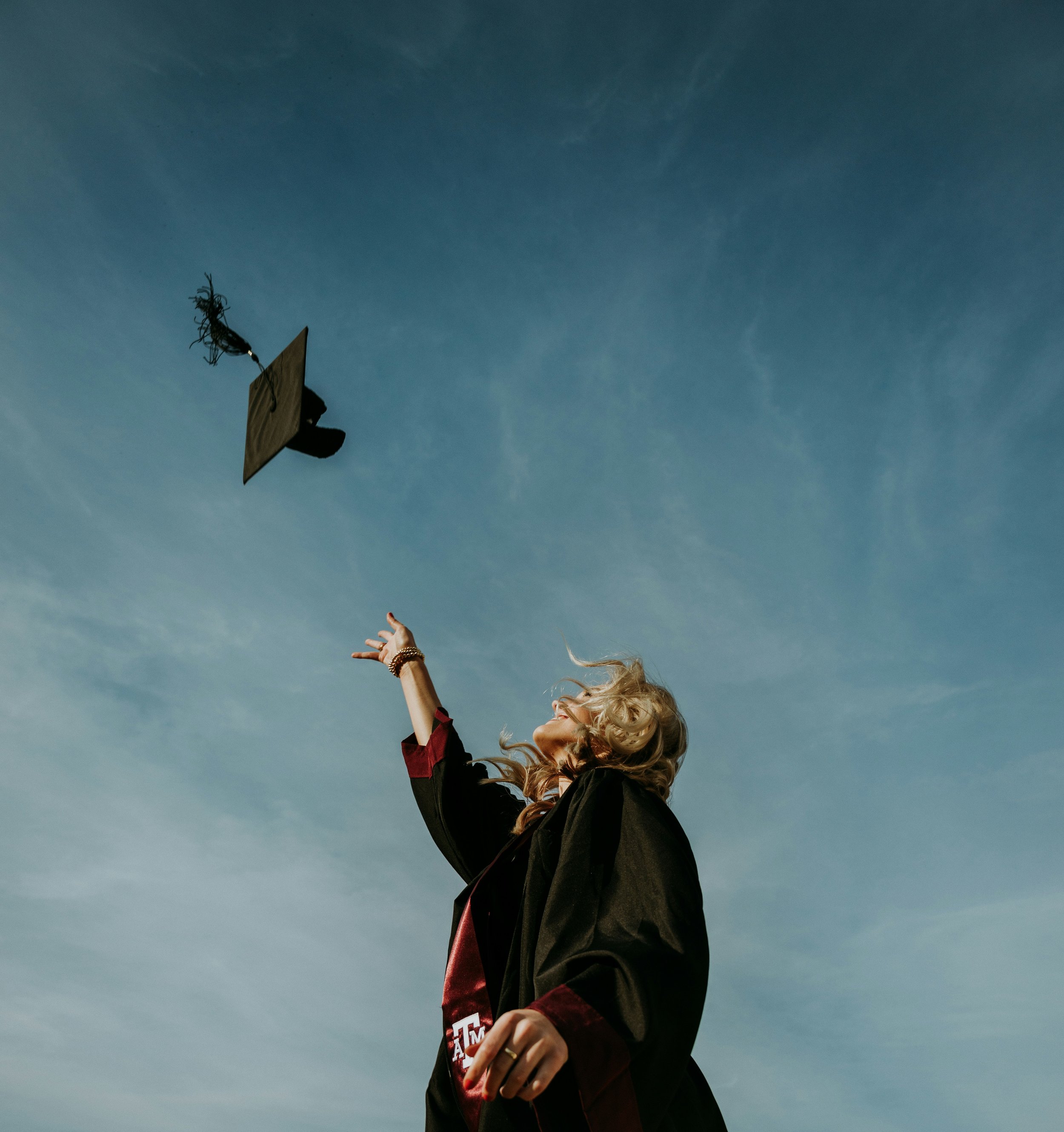 Young woman in a graduation gown tossing her cap in the air outside during daytime.