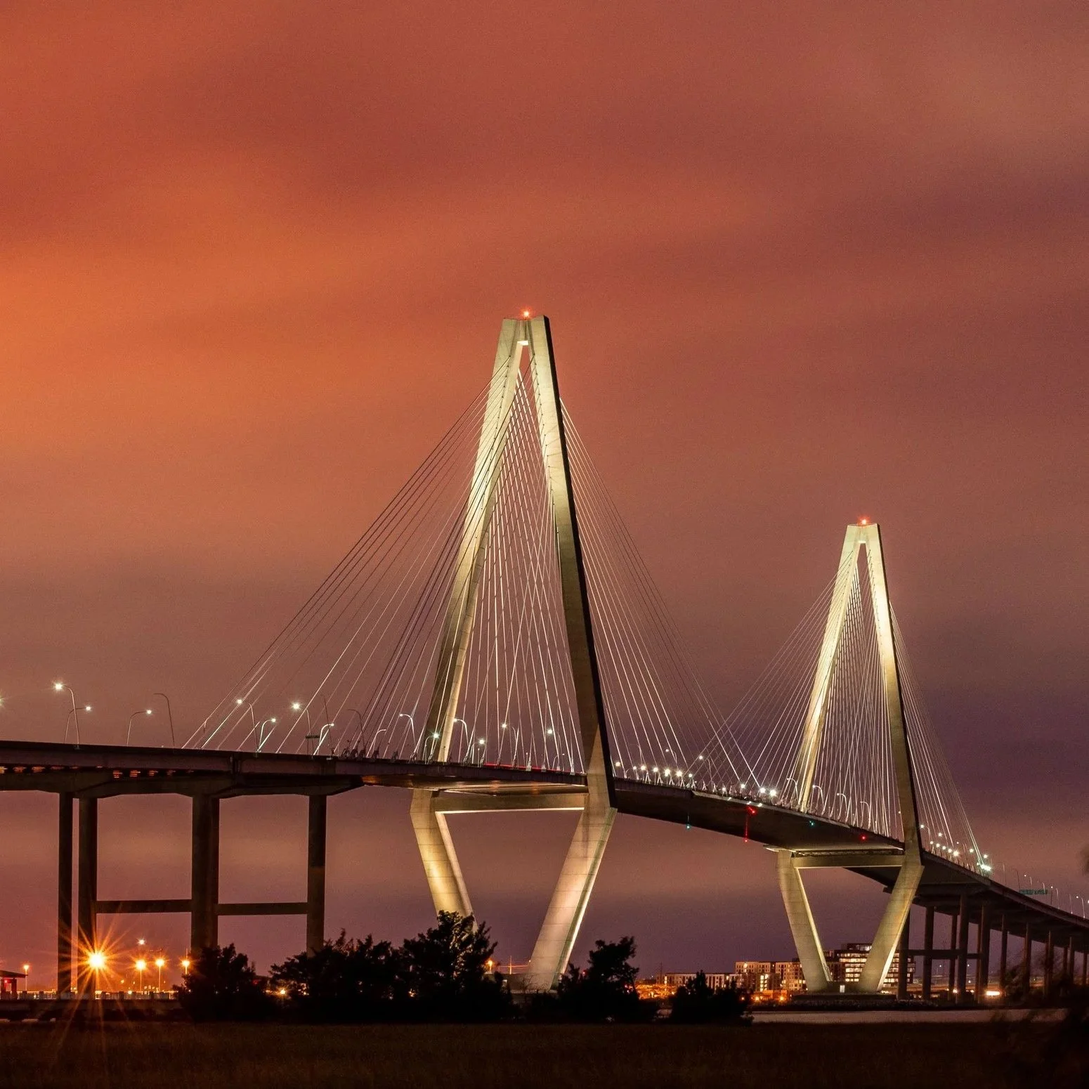 Nighttime view of the Margaret Hunt Hill Bridge in Dallas, Texas, with its white arch and cable-stayed design, illuminated against a reddish-orange cloudy sky.