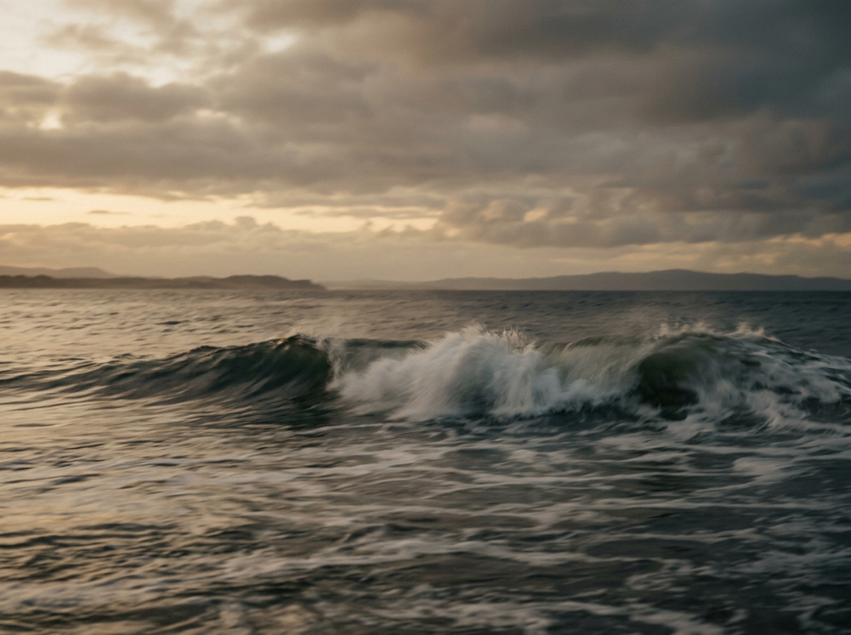 Photo of ocean waves at sunset with cloudy sky and distant landforms.