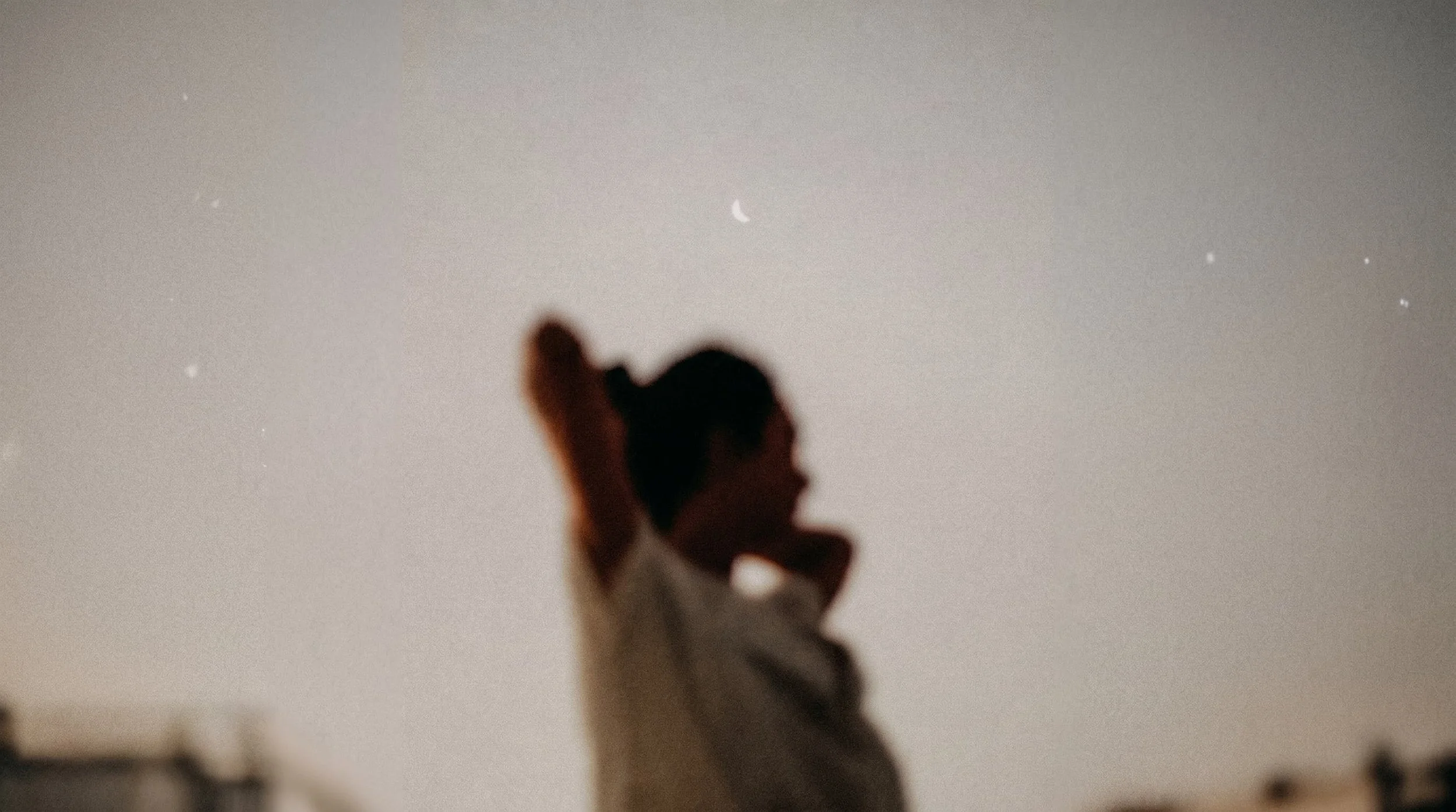 A silhouette of a person holding a heart-shaped object up in front of the night sky with a visible crescent moon and stars.