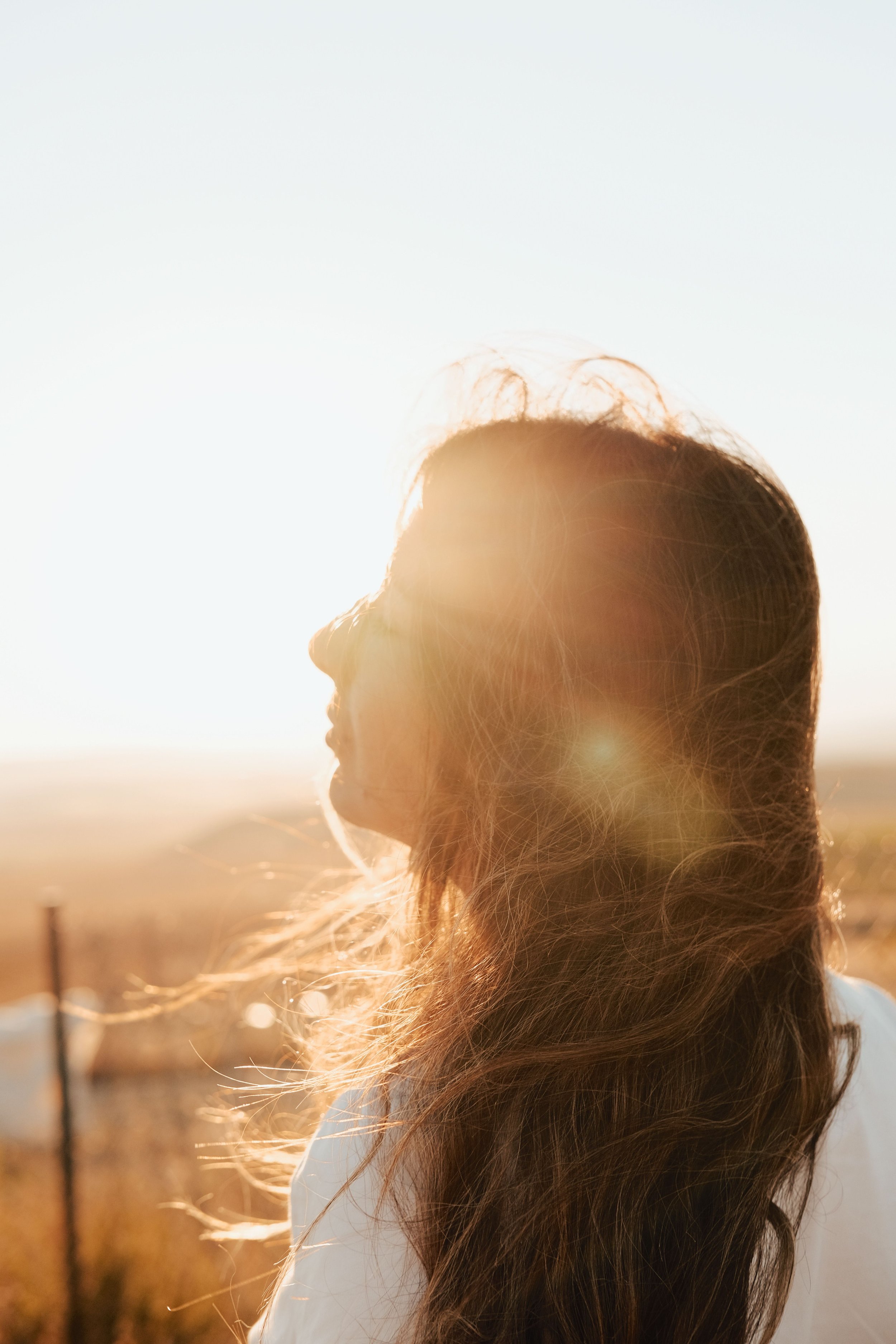 Profile of a woman with long brown hair illuminated by sunlight, with a blurred outdoor background.