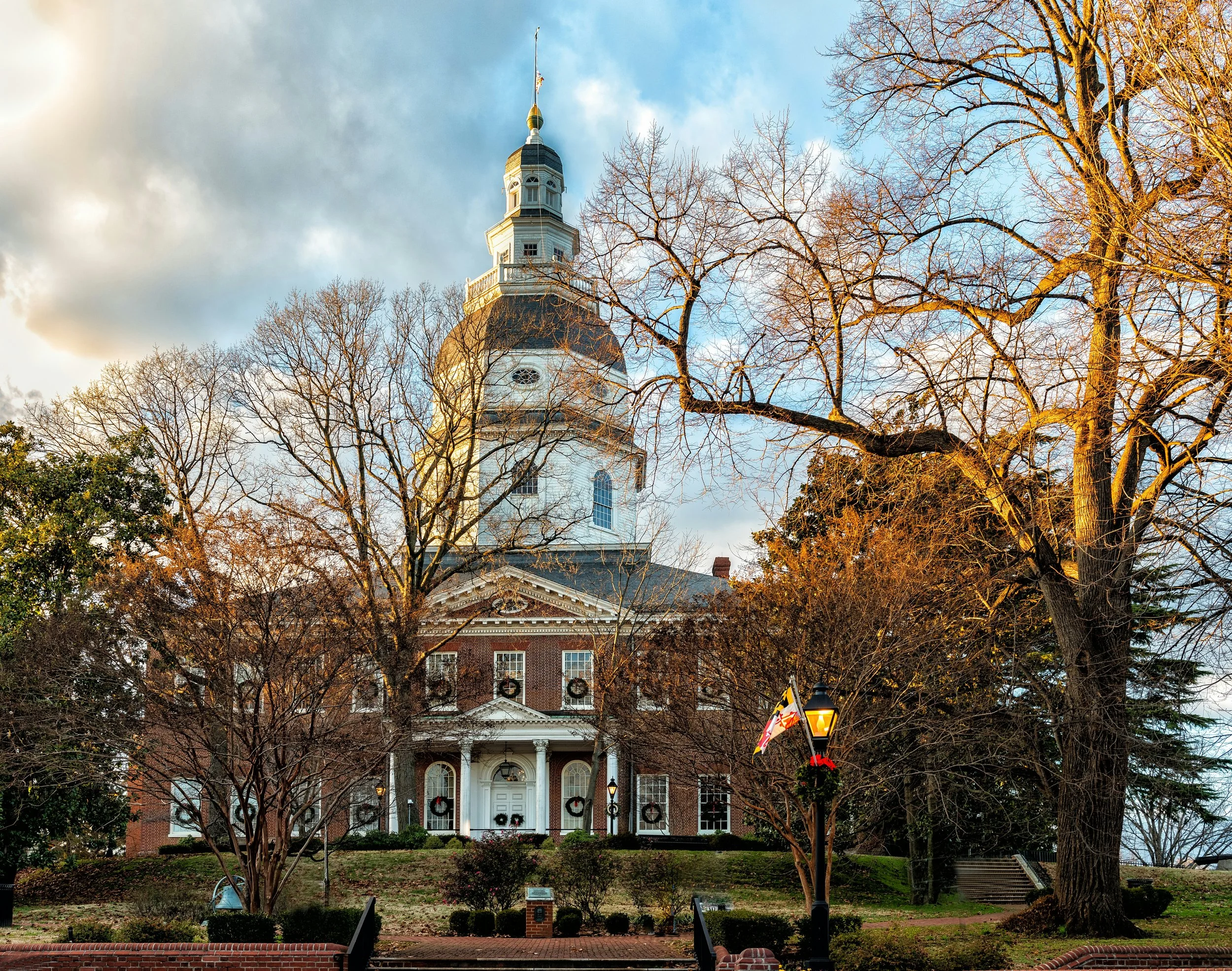 A historic brick building with white columns and a clock tower, decorated with Christmas wreaths, decorated with Christmas wreaths, and a flag flying from the top, situated in a park with leafless trees and a lamppost with holiday decorations against a partly cloudy sky.