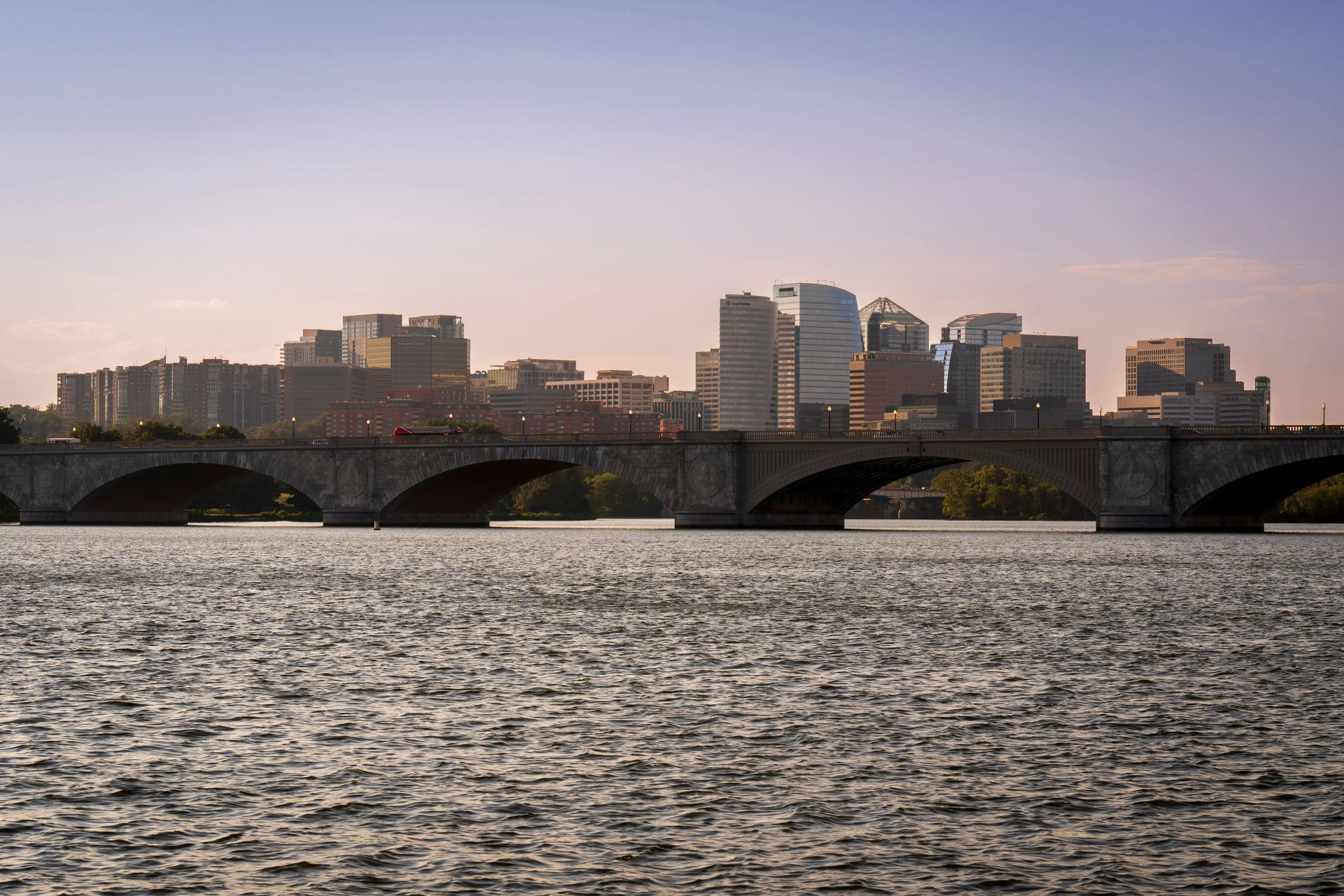 City skyline with high-rise buildings viewed across a river, with a stone bridge in the foreground and a soft pinkish sky.