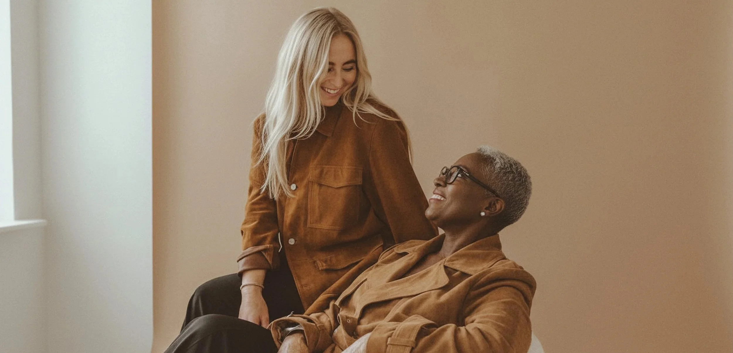 Two women with brown suede jackets sharing a moment of laughter indoors.
