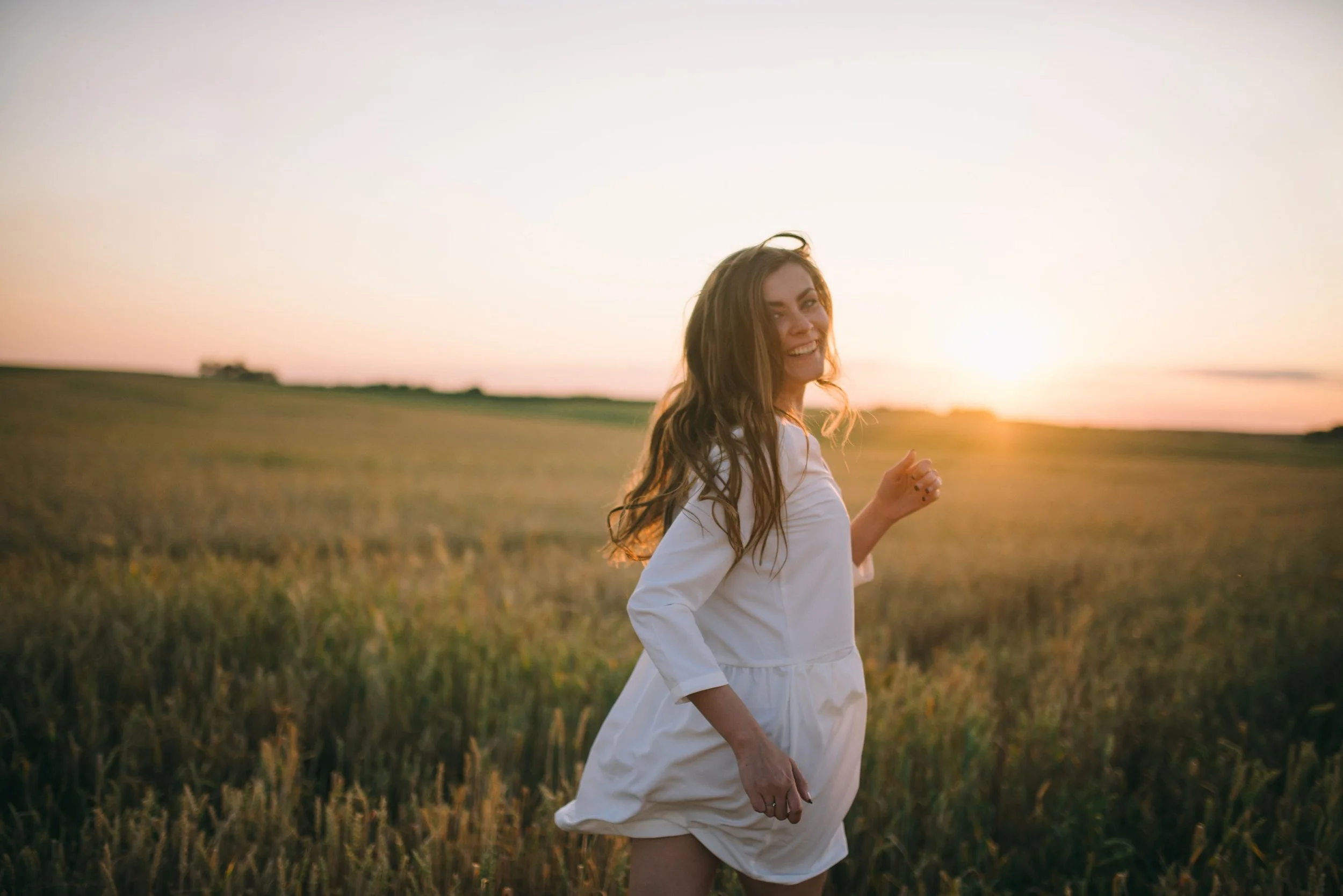 A woman with long brown hair, wearing a white dress, smiling and running in a grassy field at sunset.