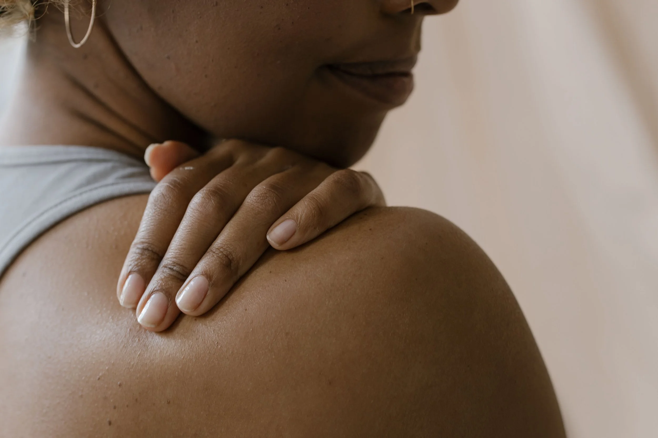 Close-up of a woman with dark skin gently holding her shoulder, wearing a white top, and a silver hoop earring visible on her left ear.