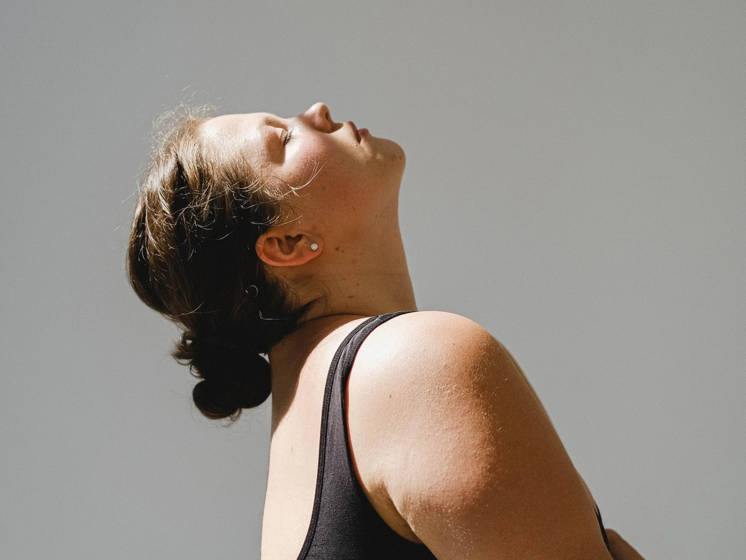 Close-up of a woman's face and shoulder with her eyes closed, head tilted back, and sunlight on her skin against a plain gray background.