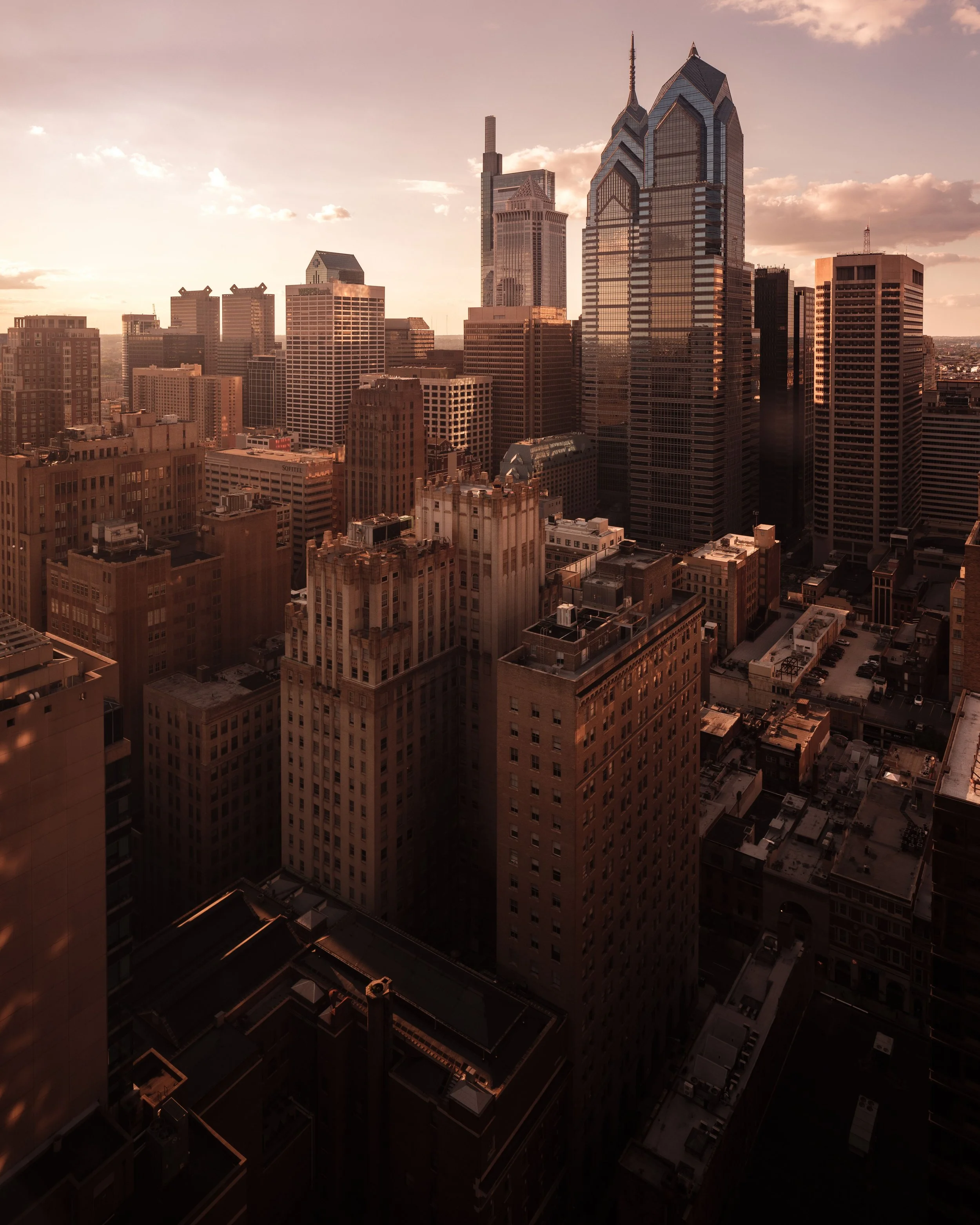 A city skyline during sunset with various skyscrapers and buildings, reflecting sunlight on glass surfaces, and partially cloudy sky.