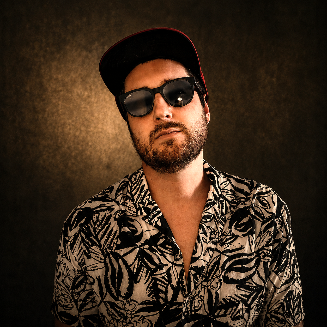 A man with a beard, wearing black sunglasses, a red and black cap, and a black and white patterned shirt, against a dark background.