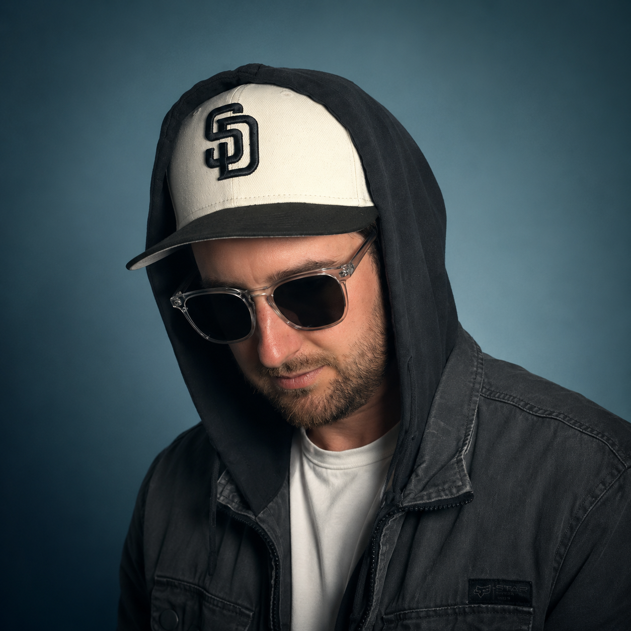 A man wearing a black hoodie, a white baseball cap with a black logo, and sunglasses, looking downward with a serious expression against a plain blue background.