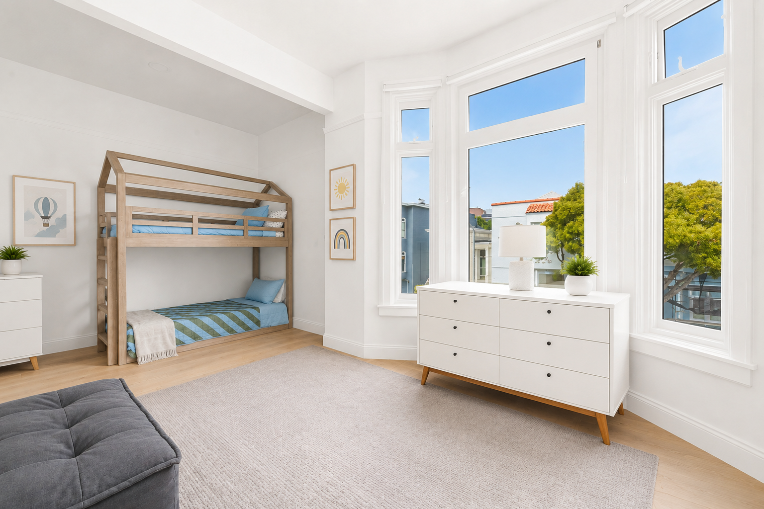 A bright, airy bedroom with white walls and large bay windows, a wooden bunk bed with blue bedding, a white dresser with a lamp and plant, and framed artwork on the walls.