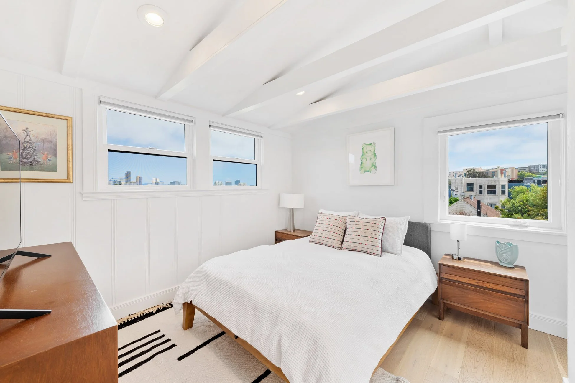 Bright bedroom with white walls and ceiling, featuring three large windows showing a cityscape with buildings, trees, and blue sky. The bed has white bedding and two striped pillows, flanked by wooden nightstands with lamps and decorative items. Artw