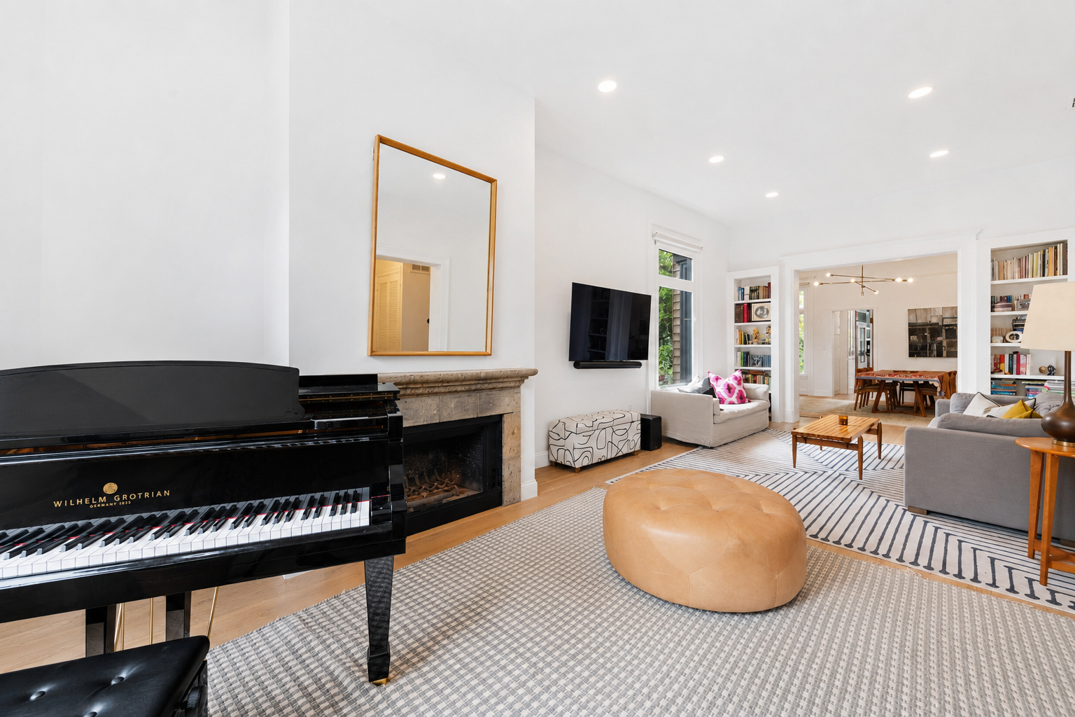 Living room with grand piano, fireplace, wall-mounted TV, white and beige sofas, area rugs, and bookshelves, with a dining area in the background.