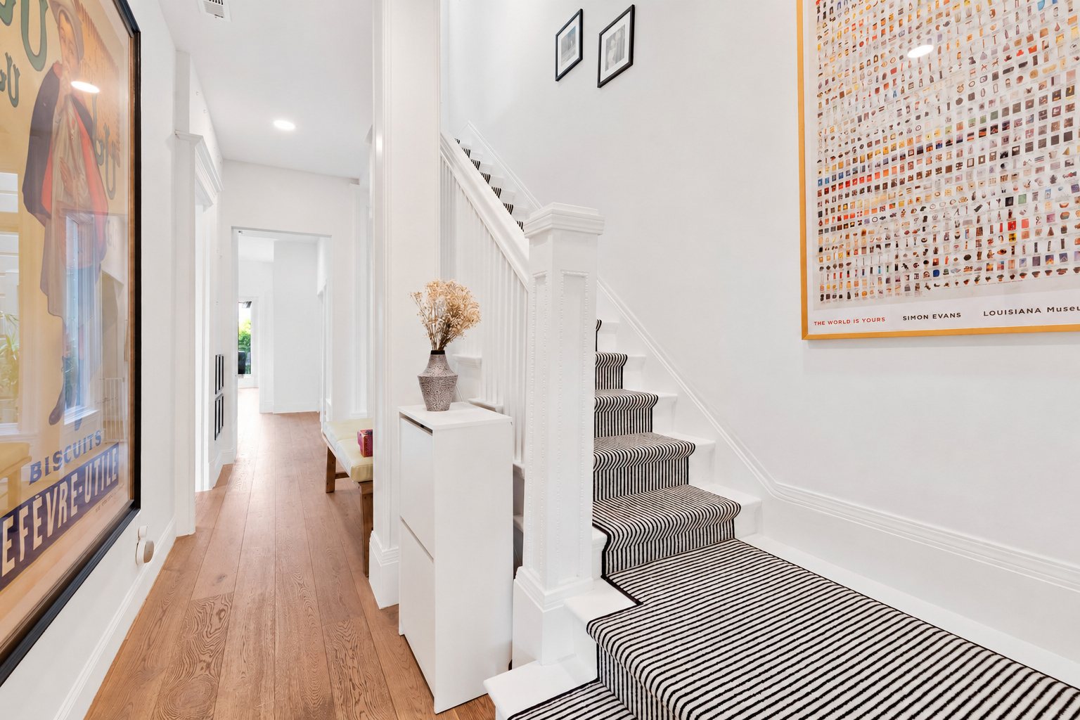 A bright interior hallway with white walls and a wooden floor. A staircase with a black-and-white striped runner leads upstairs along the right wall. There are two framed pictures on the wall above the staircase, and a large colorful framed poster ha