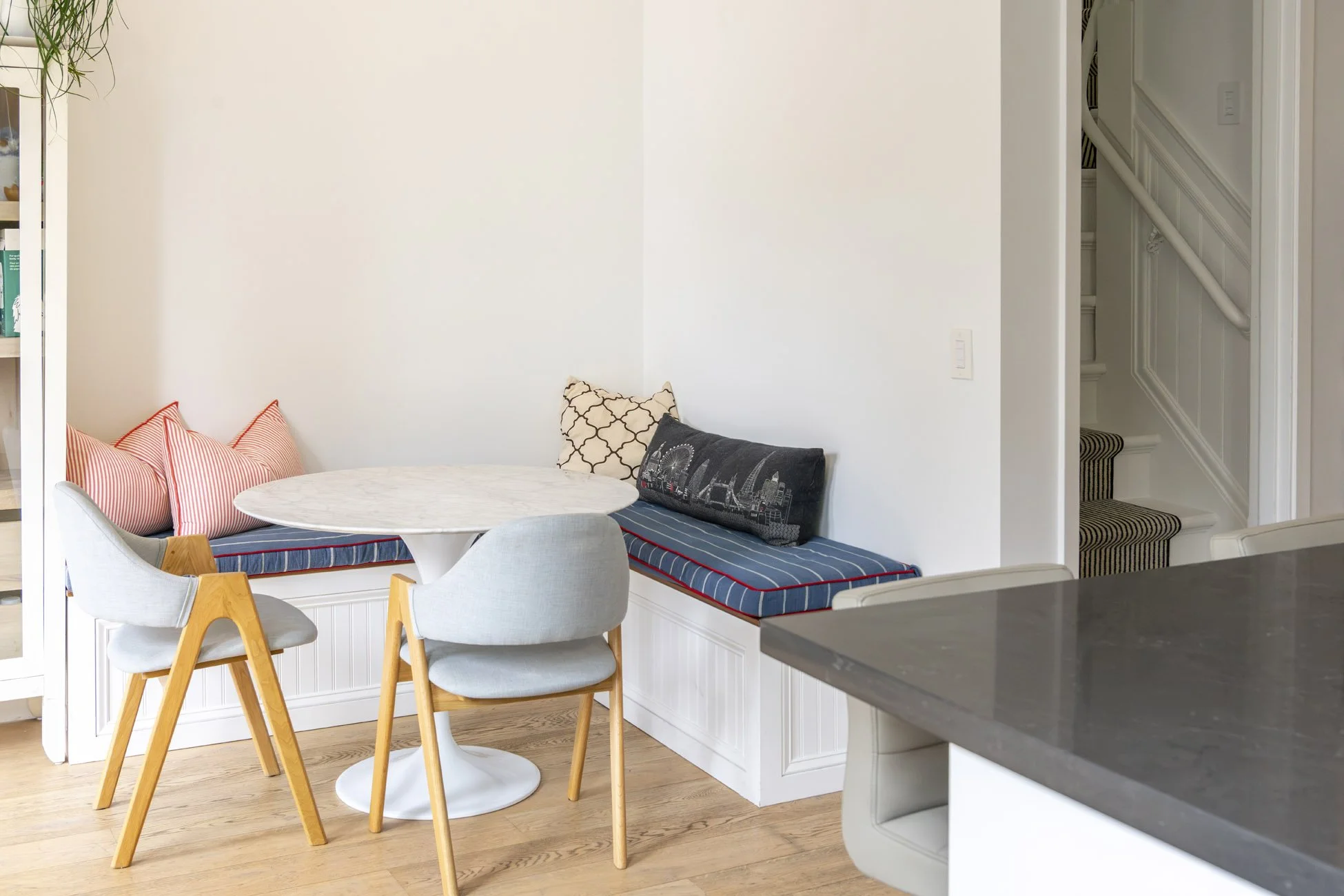 A cozy indoor sitting area featuring a white window seat with a navy blue cushion and patterned pillows including red and white stripes, beige with black geometric, and a black pillow with a cityscape design. A white round table is surrounded by a li