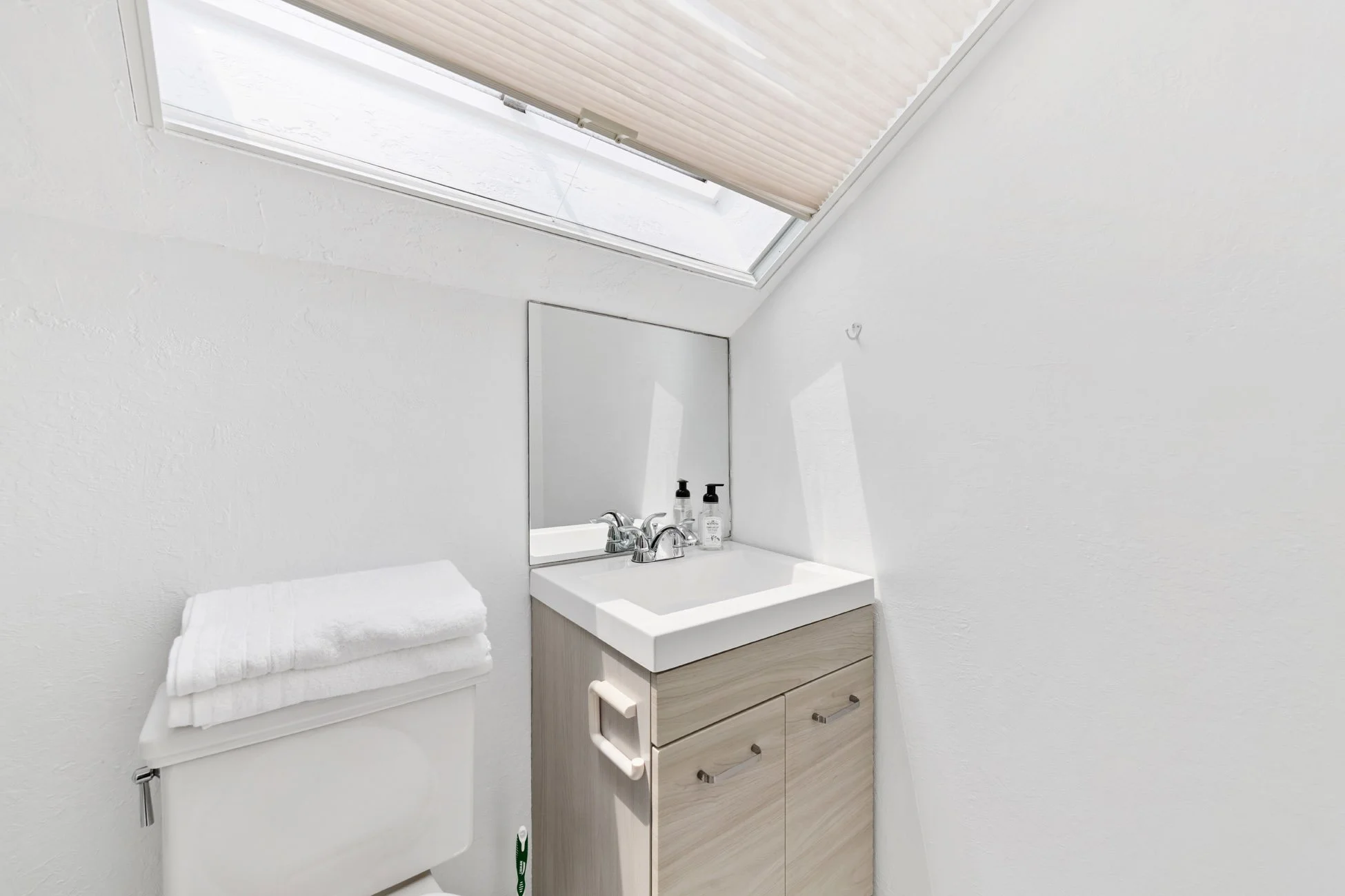 A small bathroom with a skylight window and a mirror above the sink. There is a cabinet below the sink, a towel on a small table, and soap dispensers on the sink. The walls are white and the decor is minimal.