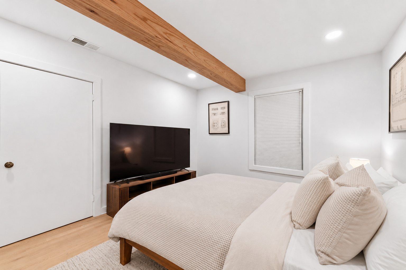 A minimalist bedroom with white walls, a large bed with white bedding and pillows, a wooden TV stand with a large flat-screen TV, a window with closed blinds, a framed picture on the wall, a wooden beam on the ceiling, and a small lamp on a bedside t
