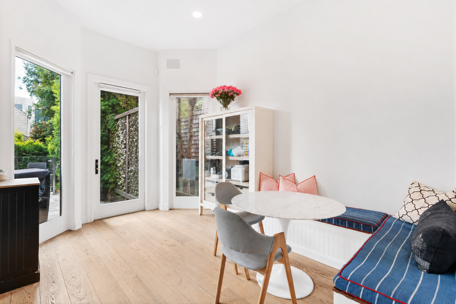 Living room with white walls, light wood floor, glass door leading outside, white cabinet with glass doors, gray chair, round table, window seat with cushions and pillows, and a vase of pink flowers on top of the cabinet.
