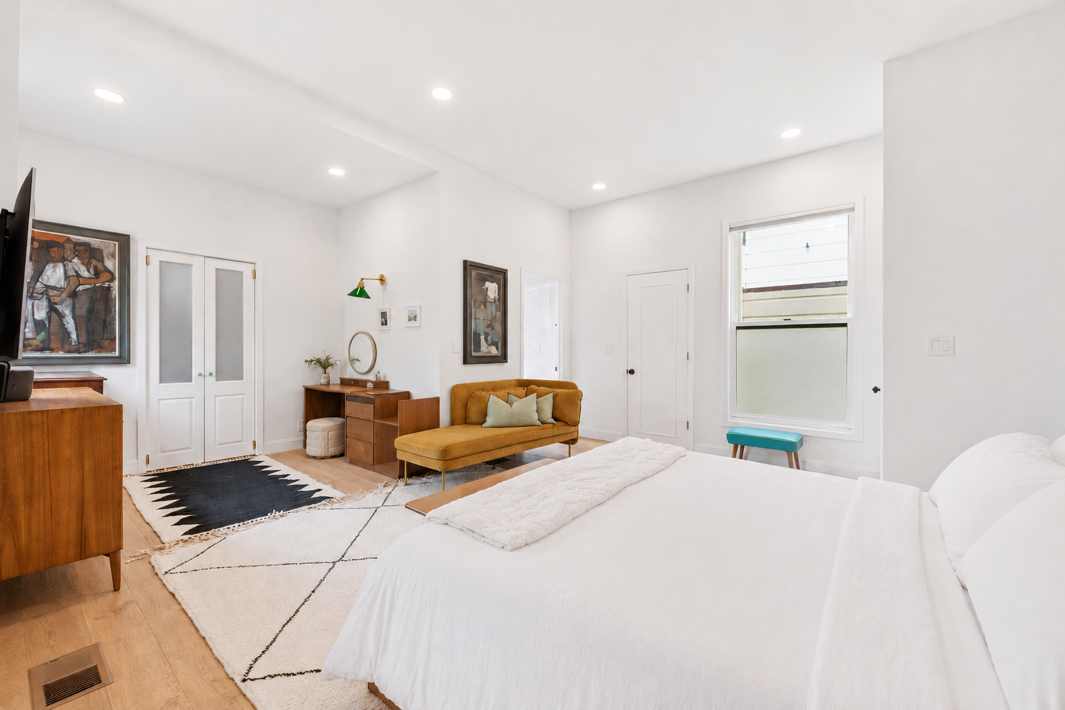Bright bedroom with white walls, a bed with white linens, a yellow velvet sofa, a wooden desk with a mirror, and a window with frosted glass.