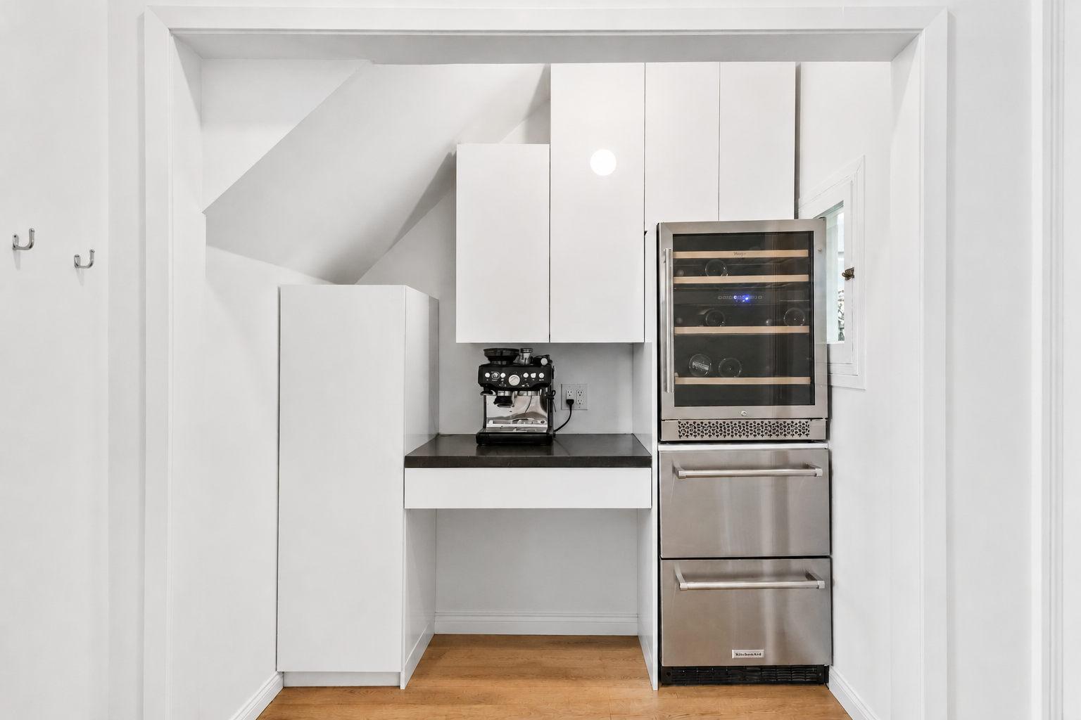 A modern kitchen nook with white cabinets, a coffee machine, and a stainless steel wine refrigerator, all installed under a sloped ceiling with wood flooring.