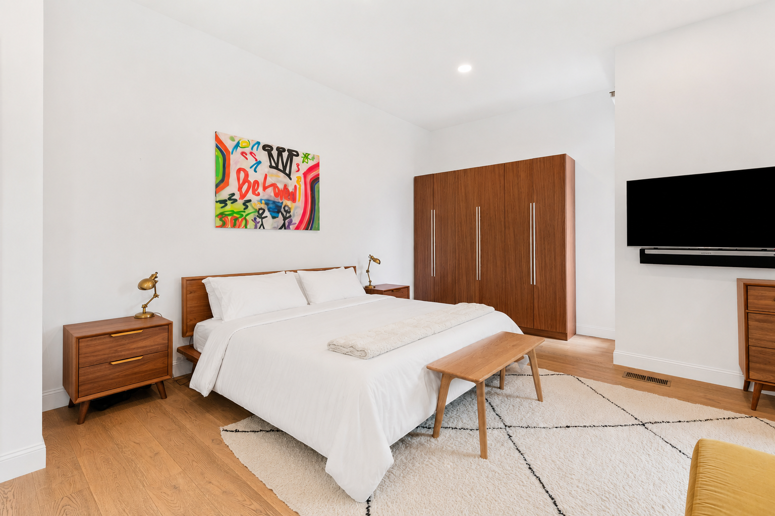 A modern bedroom with a wooden bed frame and white bedding, two matching wooden nightstands with small gold lamps, a wooden bench at the foot of the bed, a large wooden wardrobe, a wall-mounted flat-screen TV, an abstract colorful painting above the 