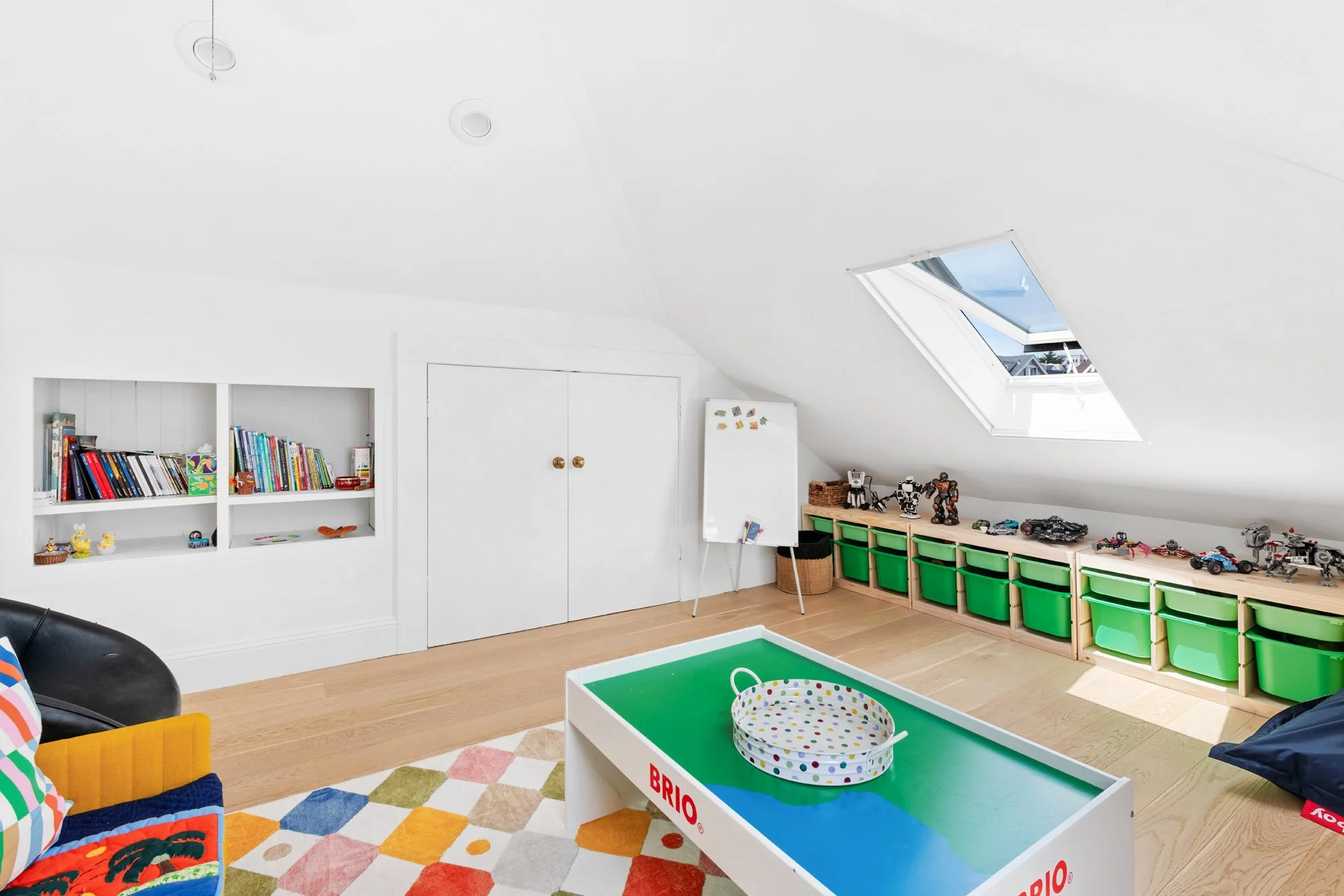 A bright playroom with white walls and sloped ceiling featuring a skylight window, with green storage bins along the right wall. On the left, a white bookshelf holds children's books and small toys. A white and green toy table with a polka dot basket