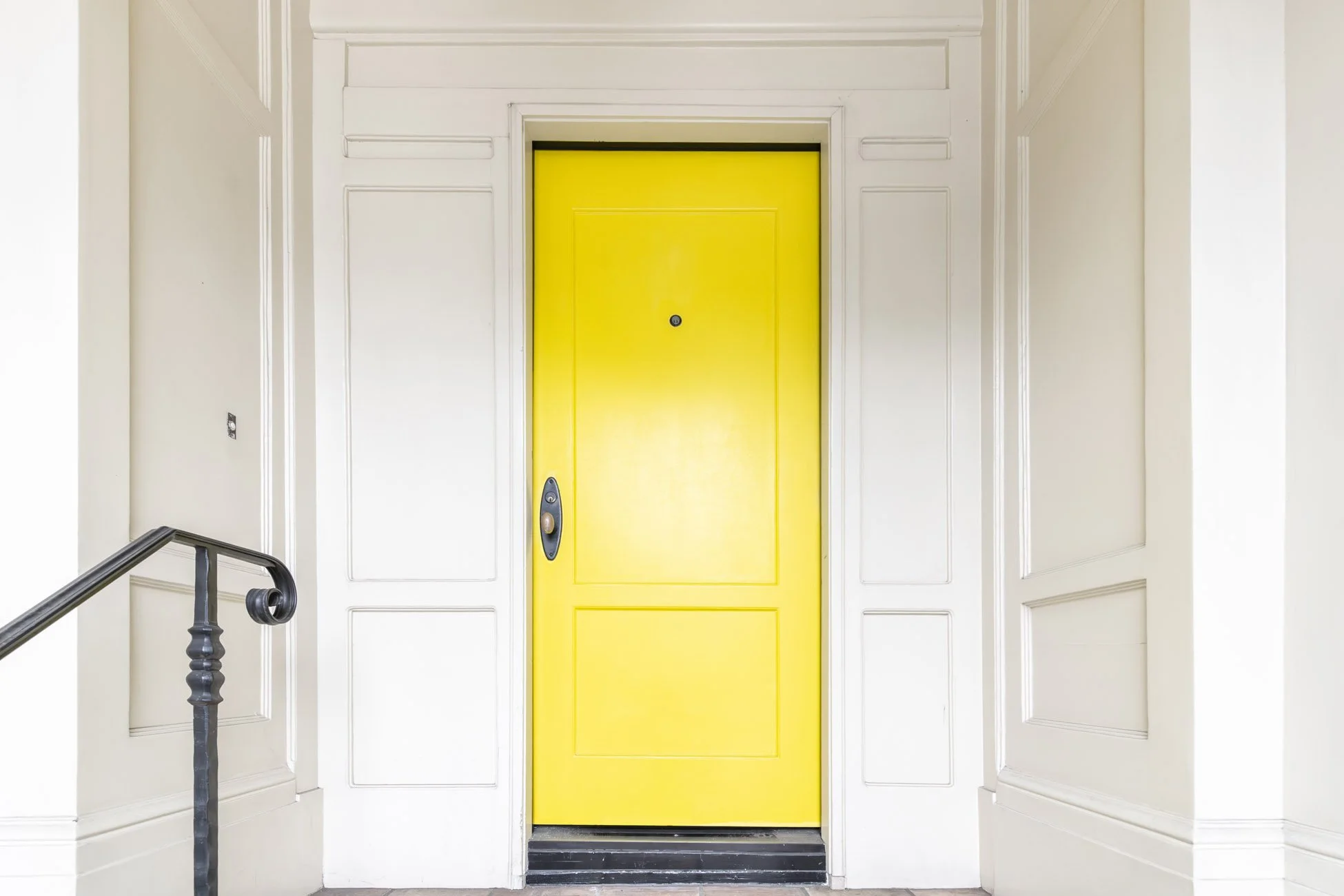 Yellow elevator door in a white wall with decorative paneling, black handle, and a doorbell in the hallway.