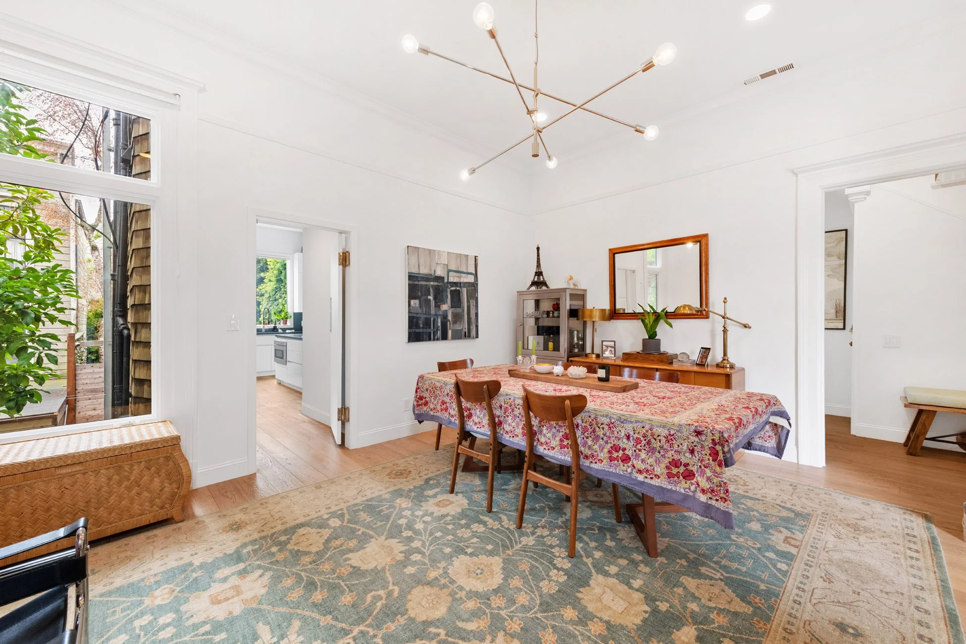 A bright dining room with a floral tablecloth-covered table, wooden chairs, a wooden sideboard, and a large mirror. An abstract painting and a small Eiffel Tower replica are on top of the sideboard. The room has white walls, hardwood floors, and a mo