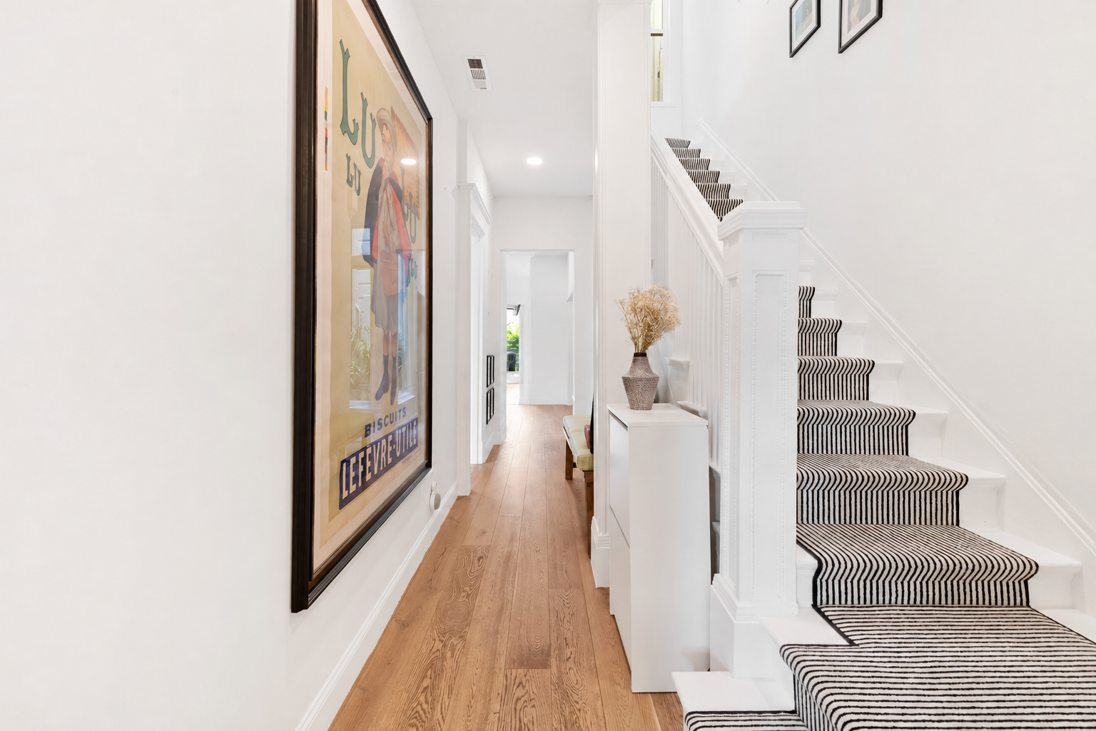 Interior view of a hallway with wooden flooring, white walls, a large framed poster on the left, a staircase with a striped black and white carpet, and a white side table with a decorative vase and dried flowers.