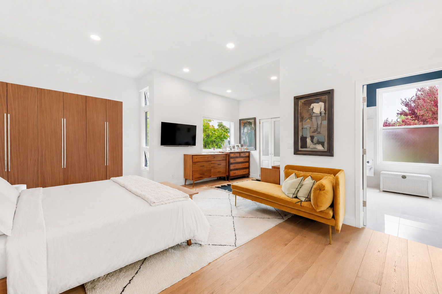 A modern bedroom with a white bed, a yellow sofa with pillows, wooden furniture, a wall-mounted TV, and large windows letting in natural light.